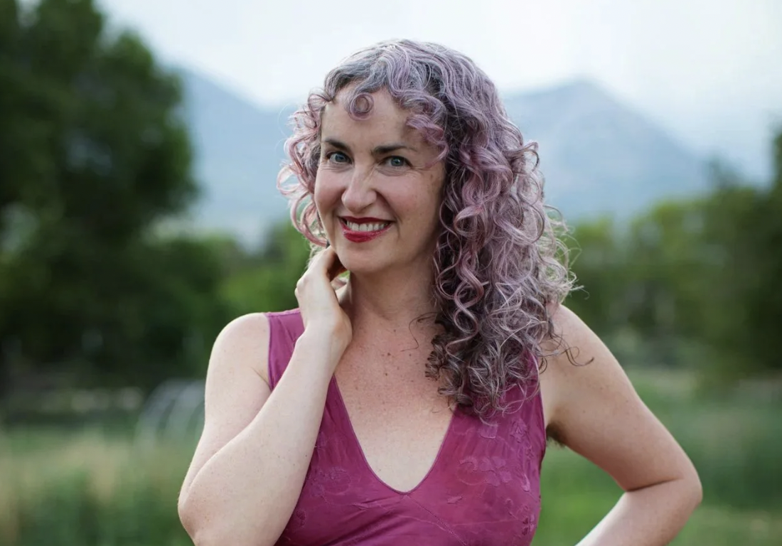 A woman with curly, purple-tinted hair outdoors, smiling, wearing a sleeveless magenta top.