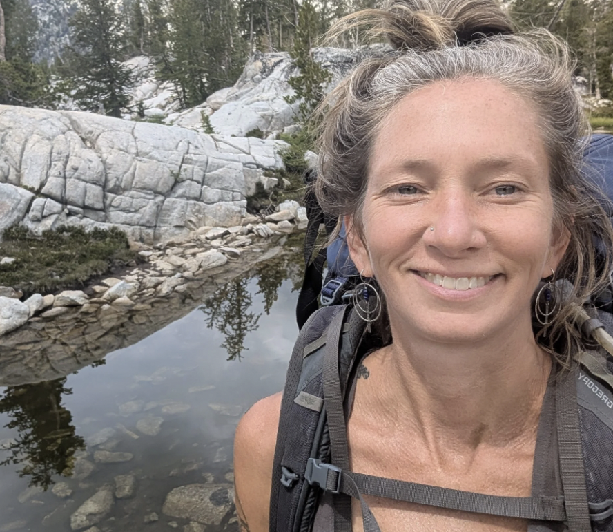 A woman with curly hair and hoop earrings smiling outdoors, carrying a backpack near a rocky mountain area with a small water body and trees.