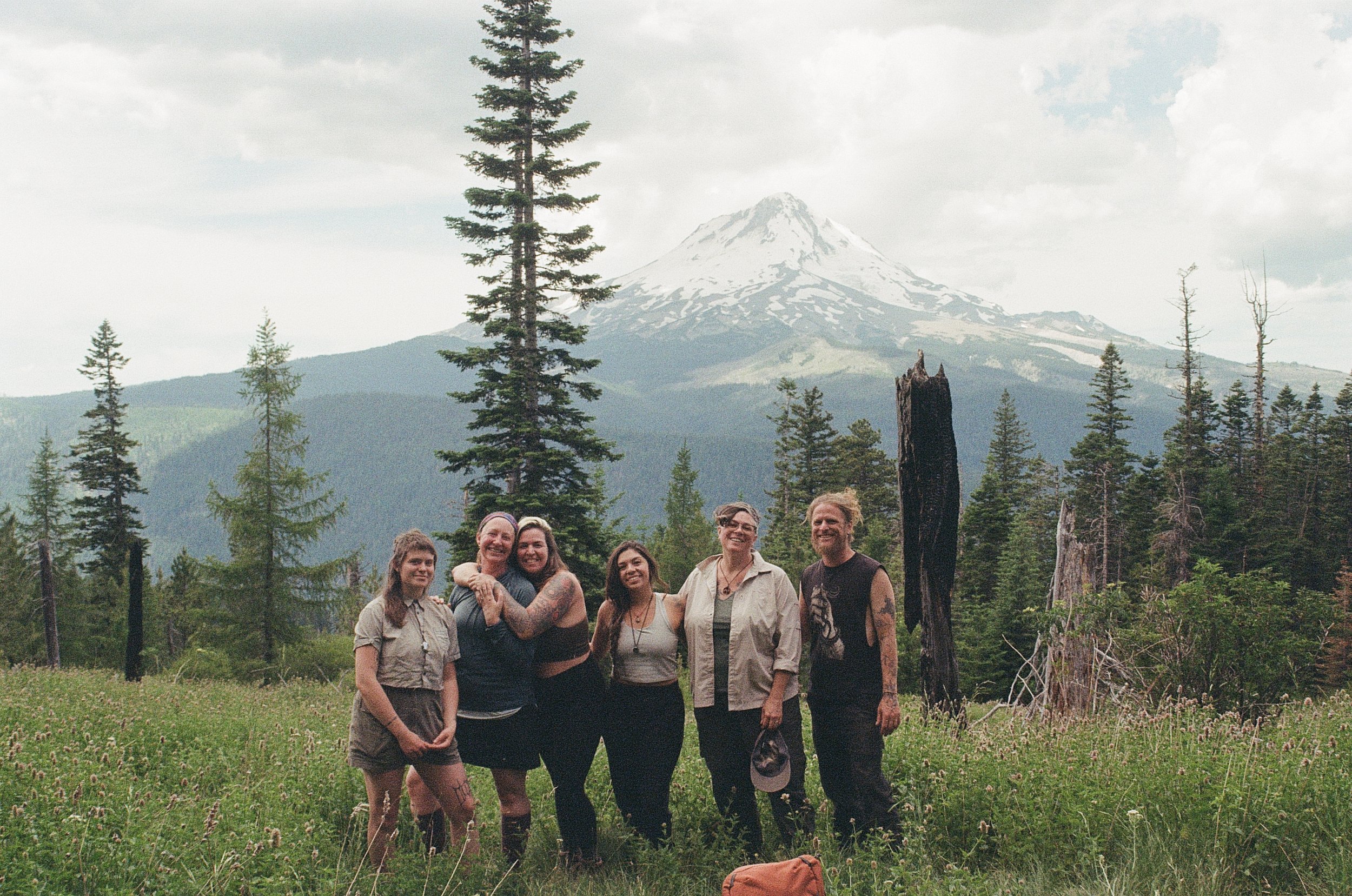 Group of eight people smiling and posing together outdoors in a forested area with Mount St. Helens in the background.