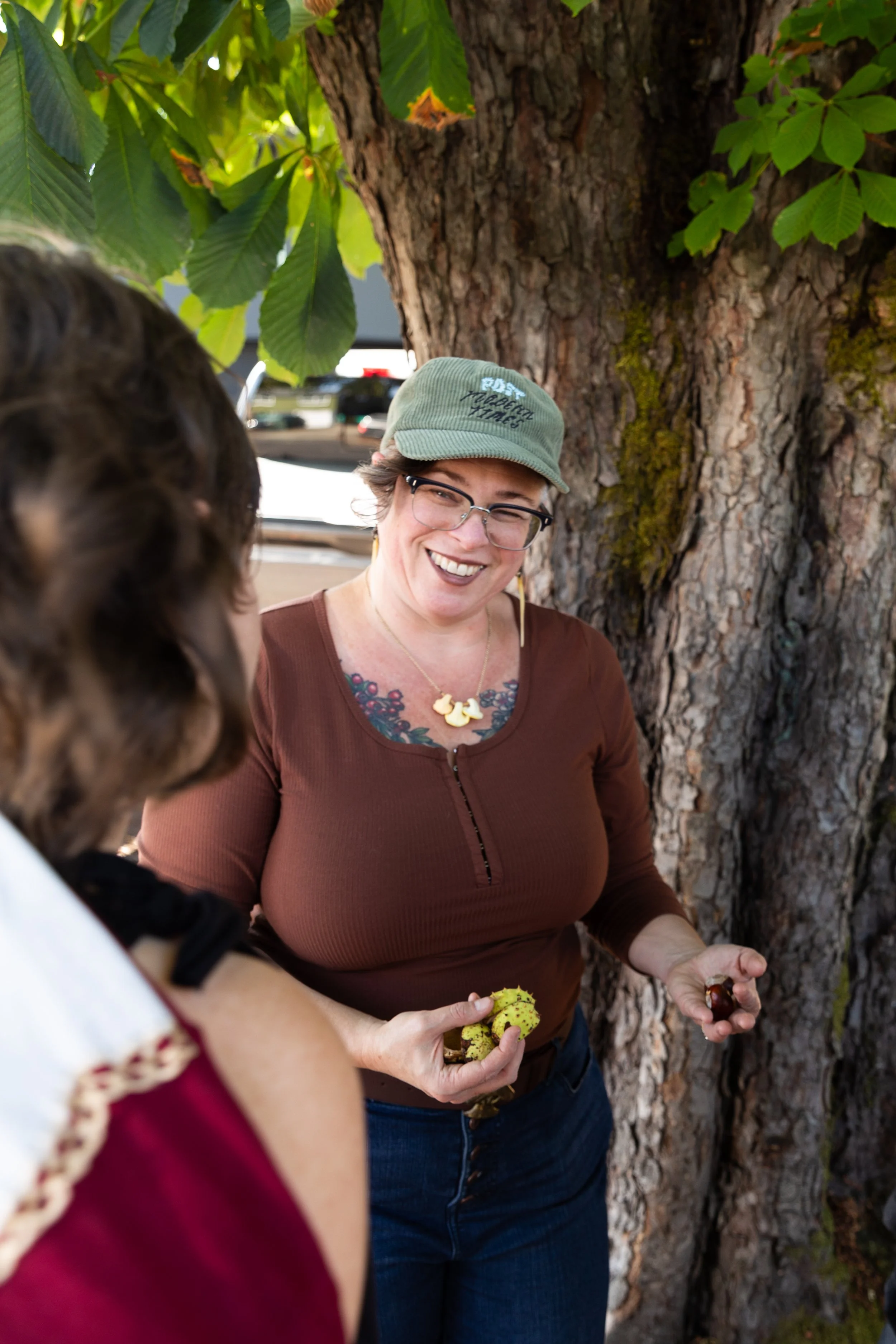 Smiling woman wearing glasses and a green cap, standing outdoors near a tree, holding yellow and green spiky objects, possibly fruits or seeds, engaging in conversation with children.