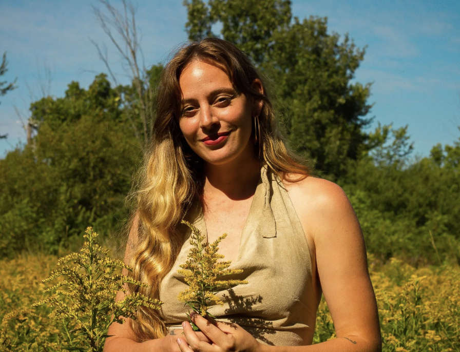 A woman with long wavy blonde hair standing in a field of yellow flowers during daytime, smiling at the camera with trees and a blue sky in the background.