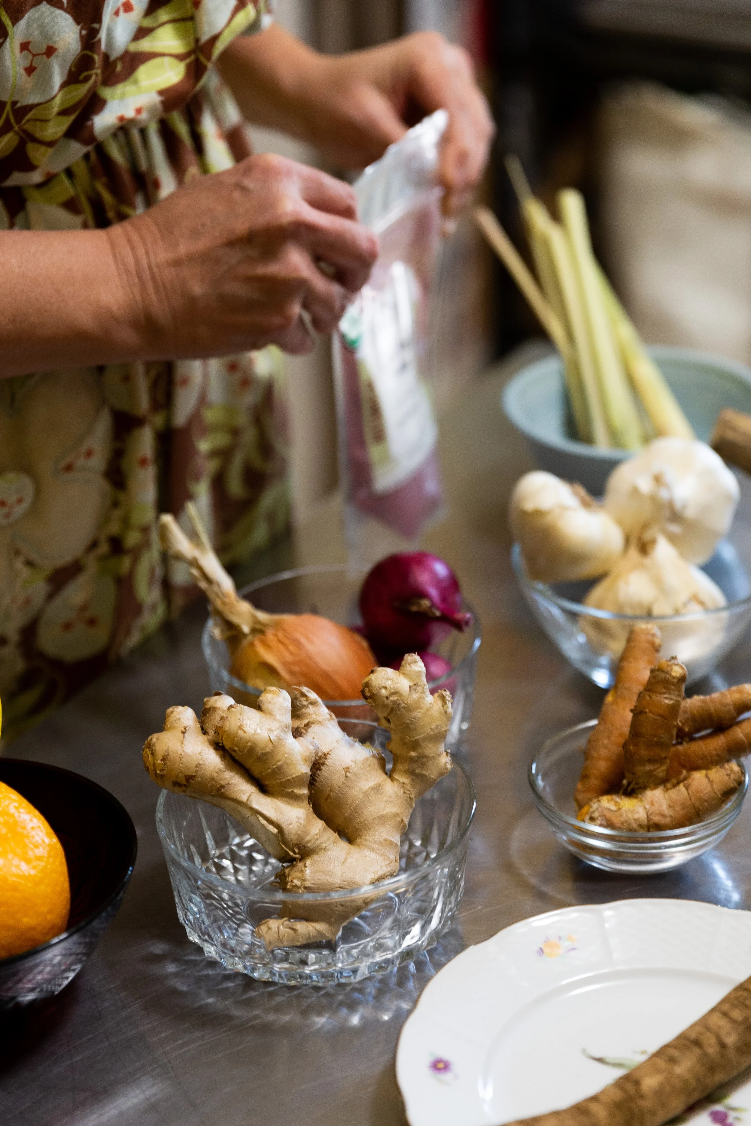 Various fresh vegetables including ginger, onions, radishes, and carrots arranged in glass bowls on a table, with a person in a patterned shirt preparing food.