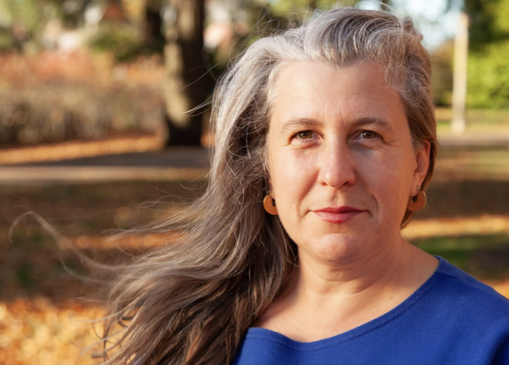 A woman with long gray hair wearing a blue shirt and orange earrings outdoors in a park with fall foliage.