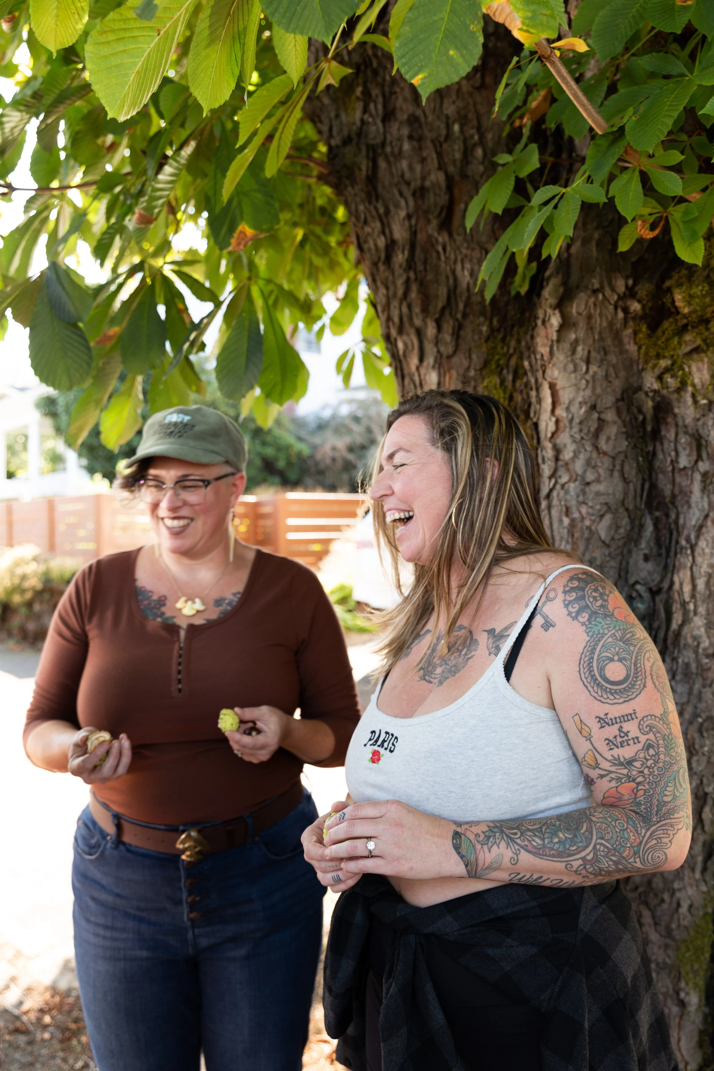 Two women standing outdoors near a large tree, smiling and laughing, holding snacks in their hands.
