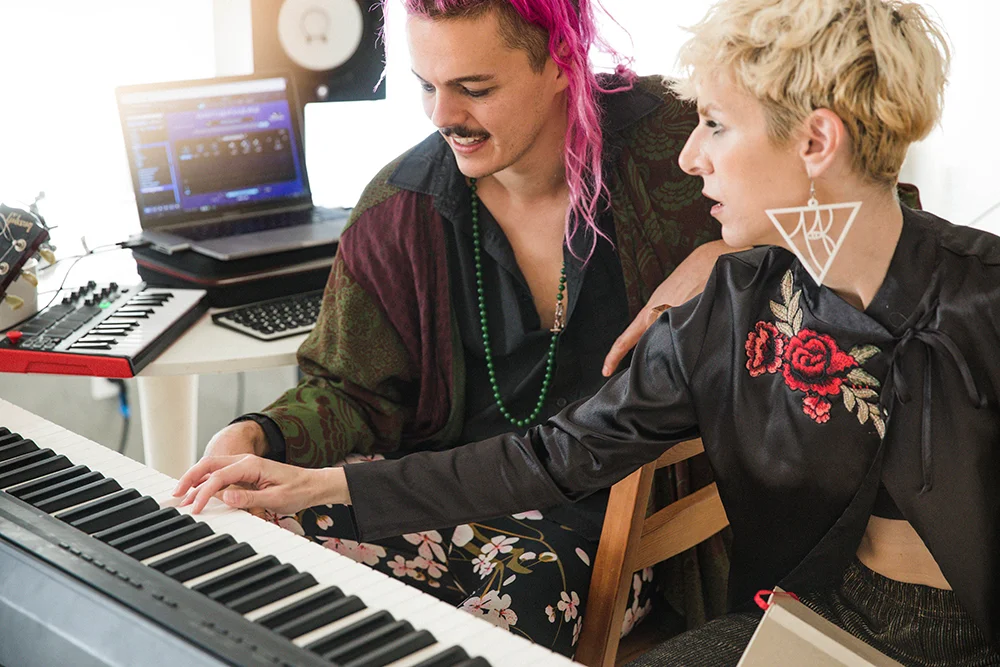 Blonde woman teaching man with pink hair how to play the piano