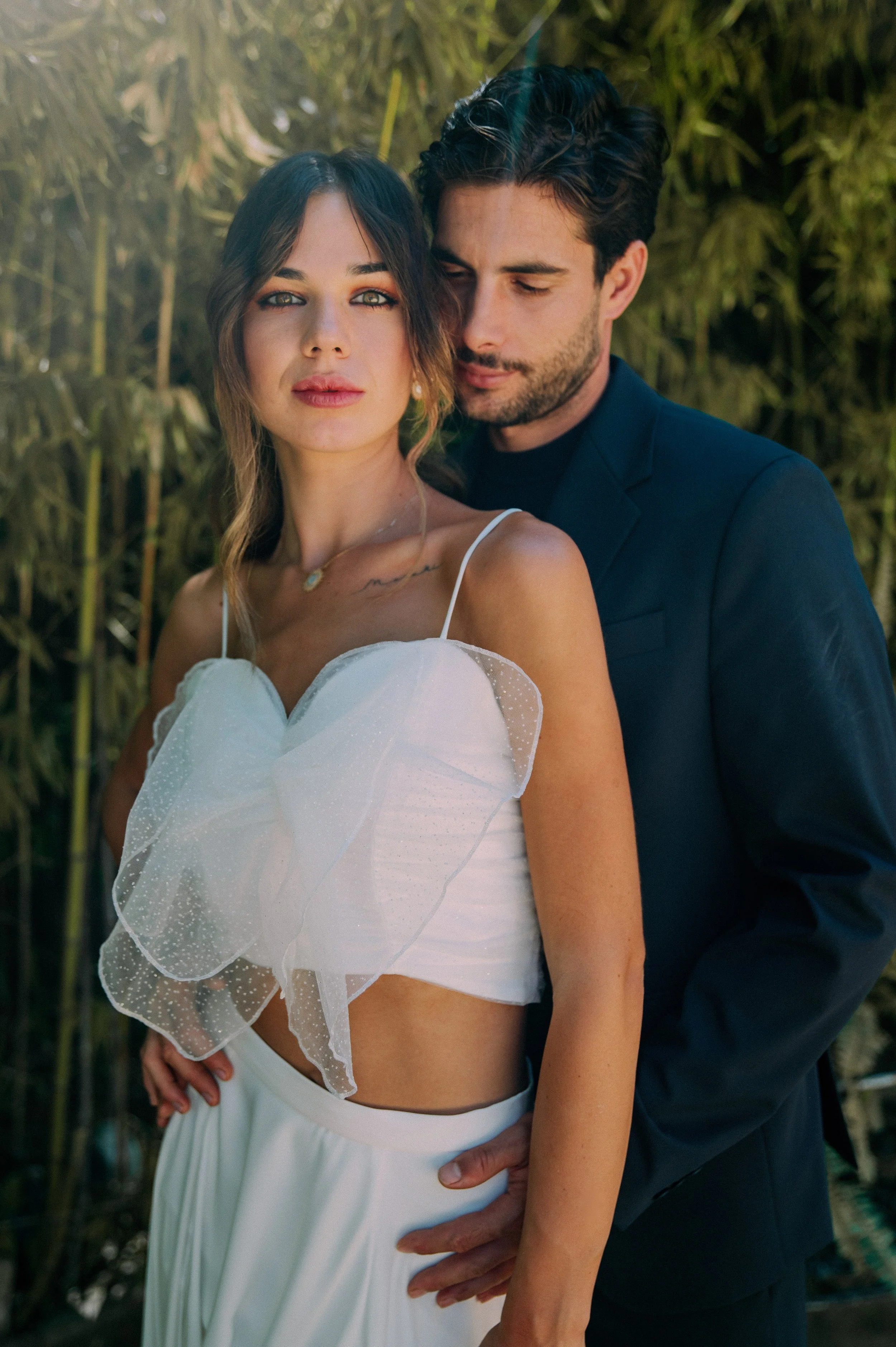 A couple standing close together outdoors, with the woman in a white dress with spaghetti straps and ruffled details, and the man in a dark suit, behind her and gently holding her waist.