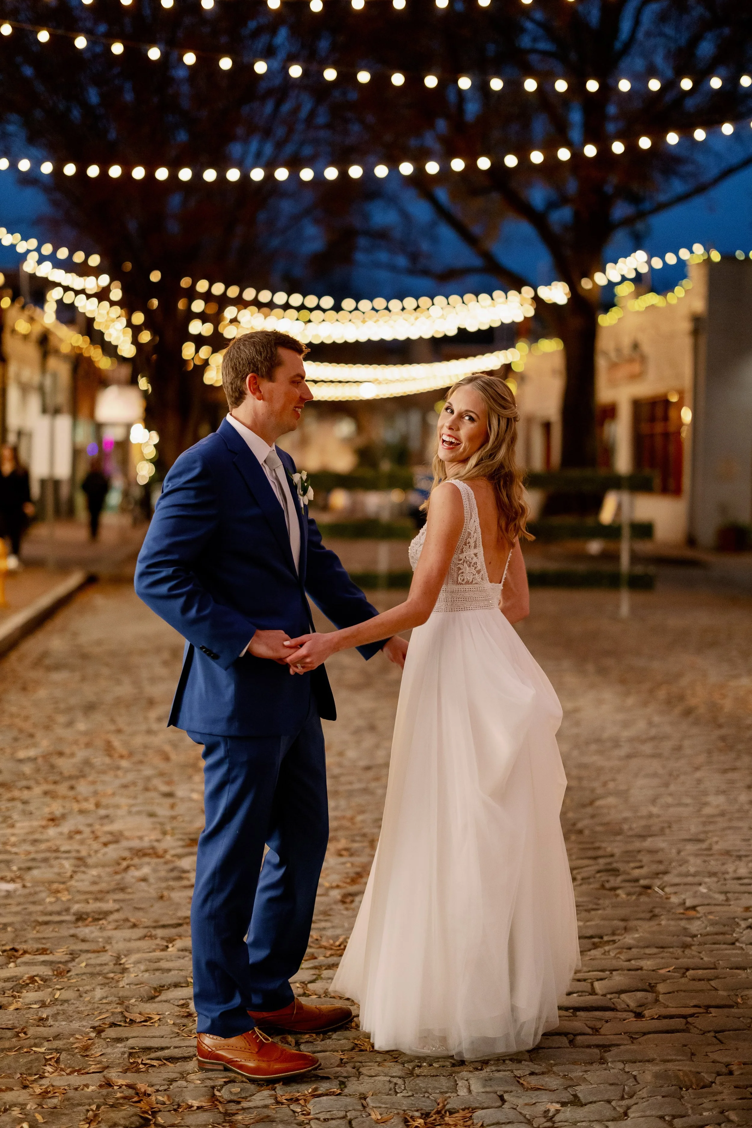 Bride and groom holding hands under string lights at Market Hall in downtown Raleigh during an evening wedding celebration