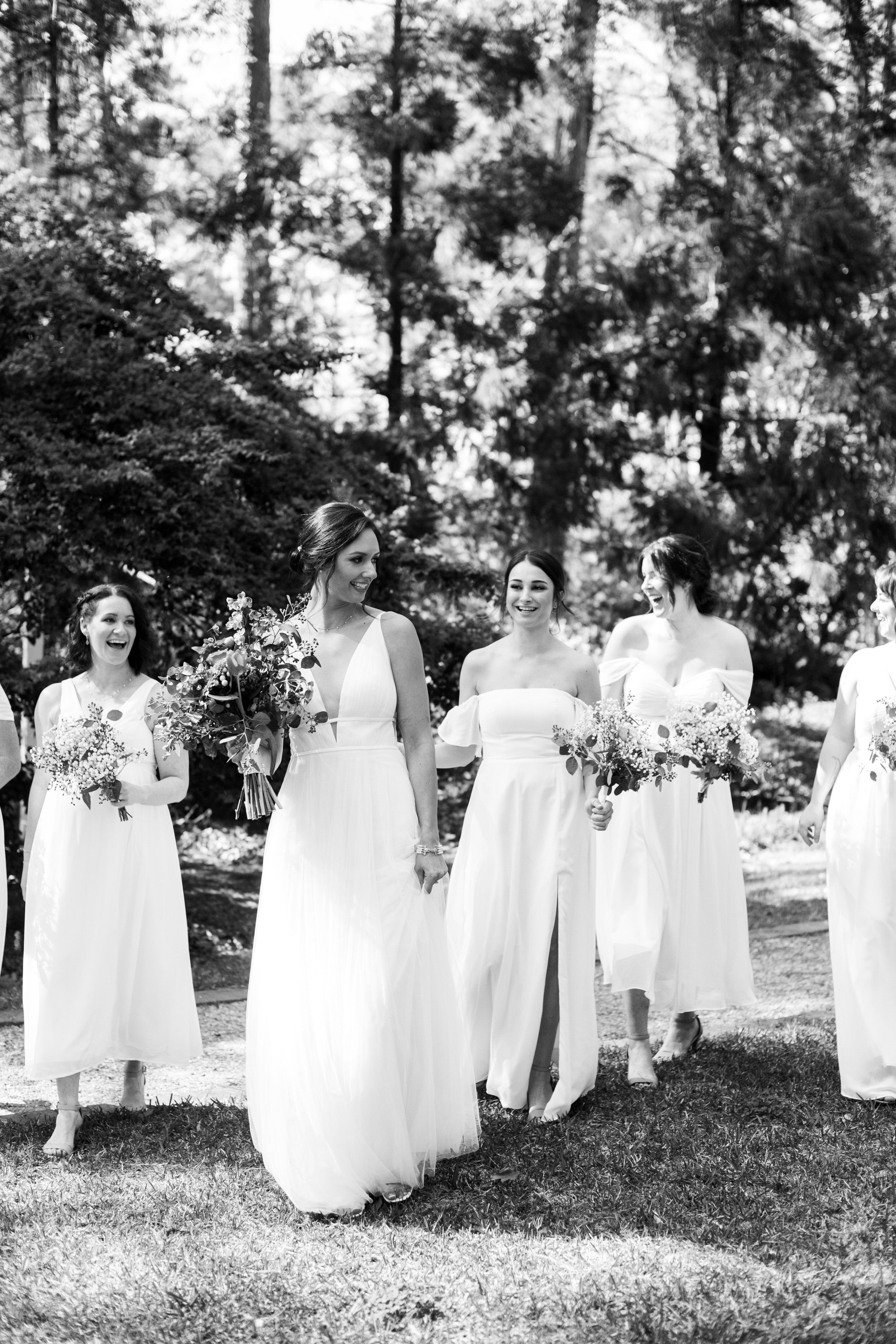  Black and white image of bride and bridesmaids in all-white dresses walking in Cape Fear Botanical Garden wedding venue.  