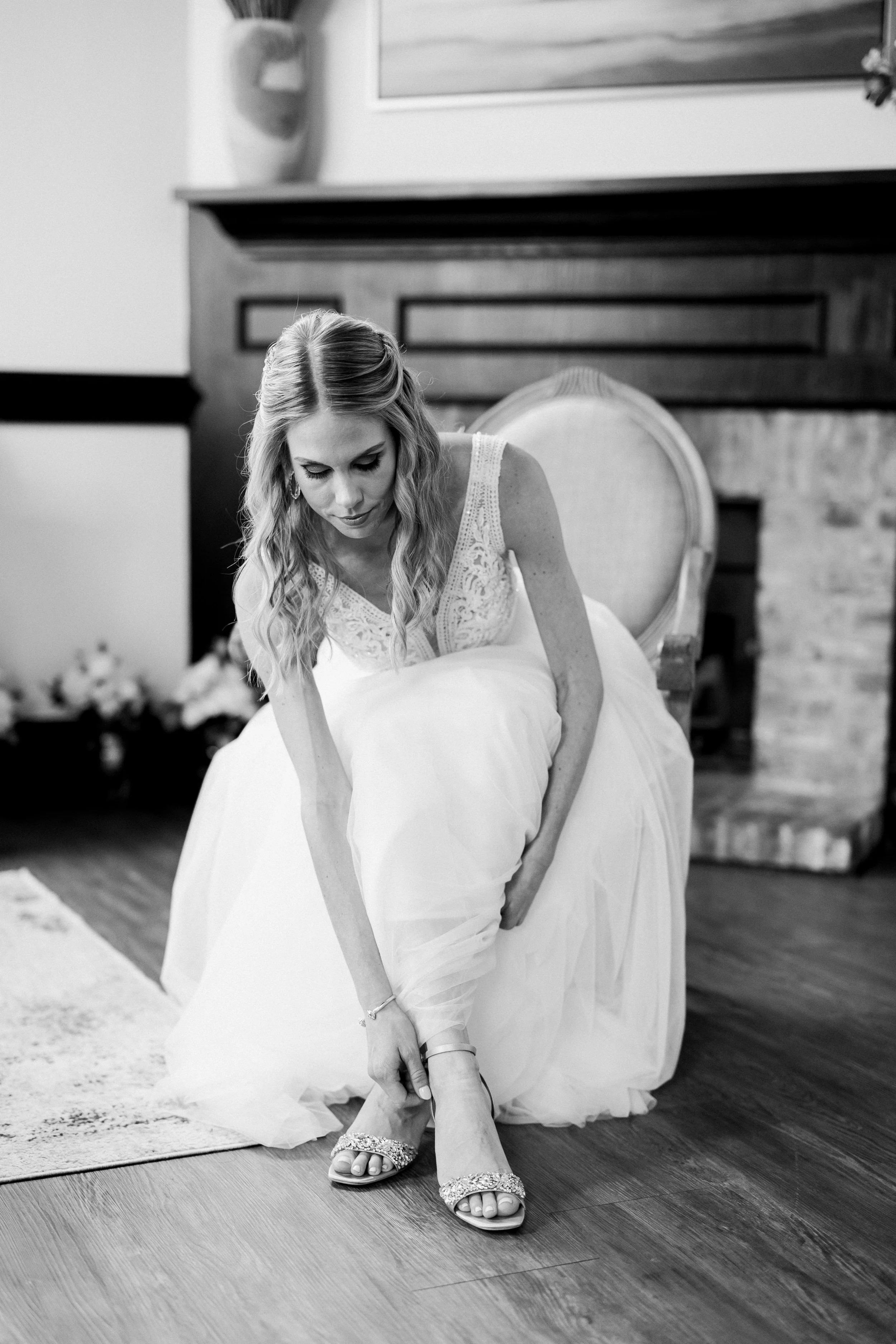 Bride sitting in a vintage chair and fastening her shoes while getting ready for her Market Hall wedding in Raleigh