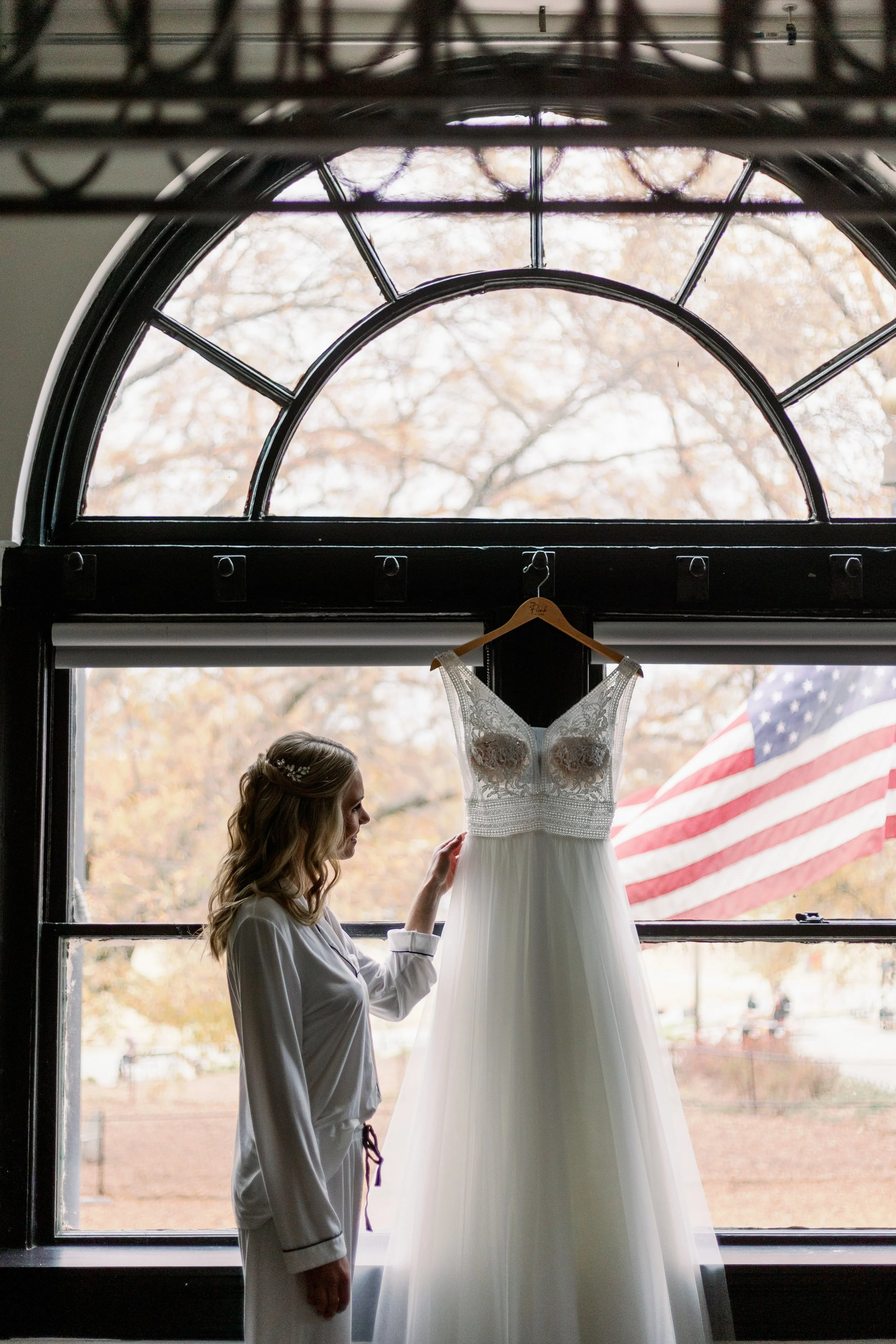 Bride in pajamas looking at her wedding dress hanging in front of a large arched window with an American flag outside at Market Hall in Raleigh