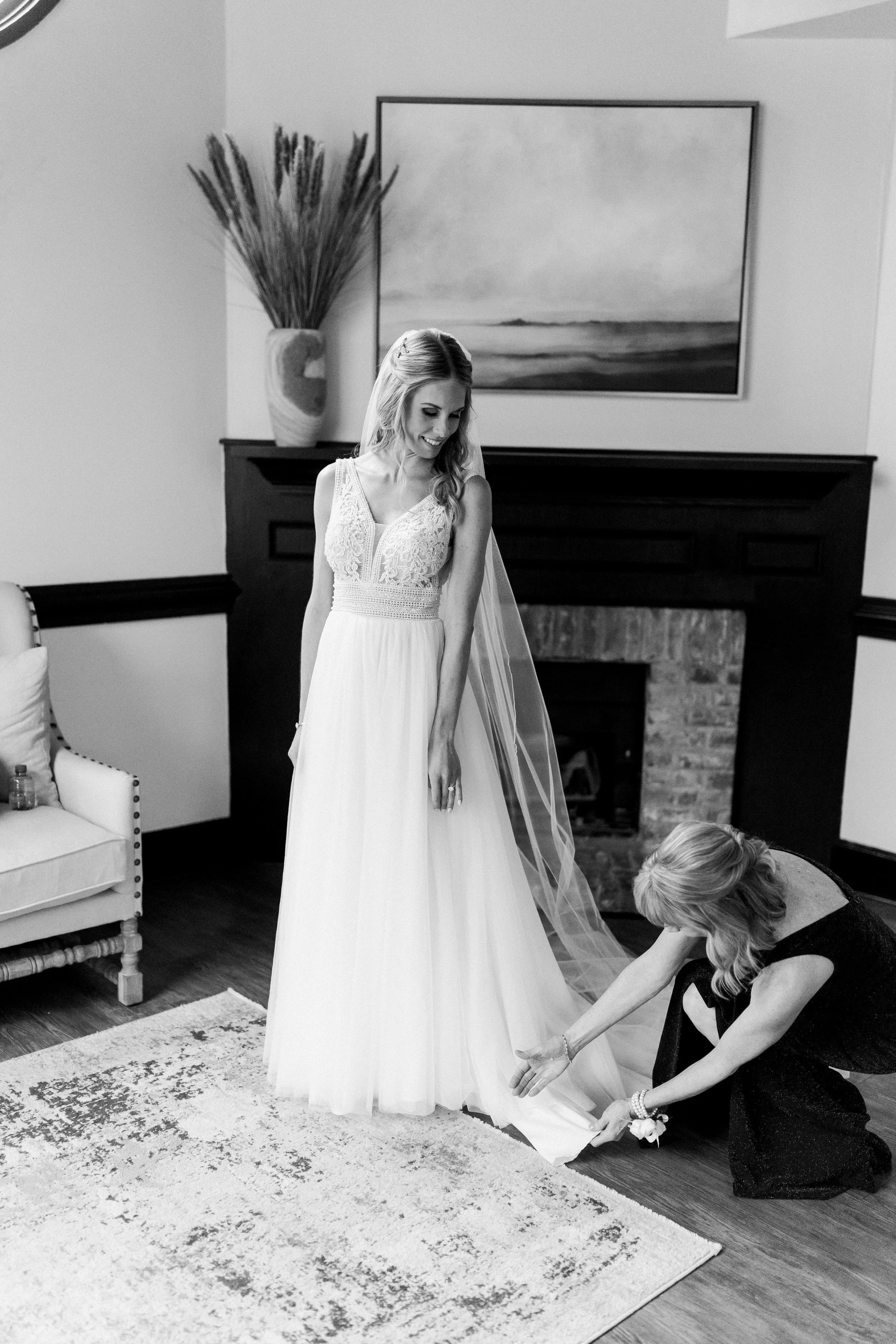 Bride smiling while her mother adjusts the hem of her gown during getting ready moments at Market Hall in Raleigh