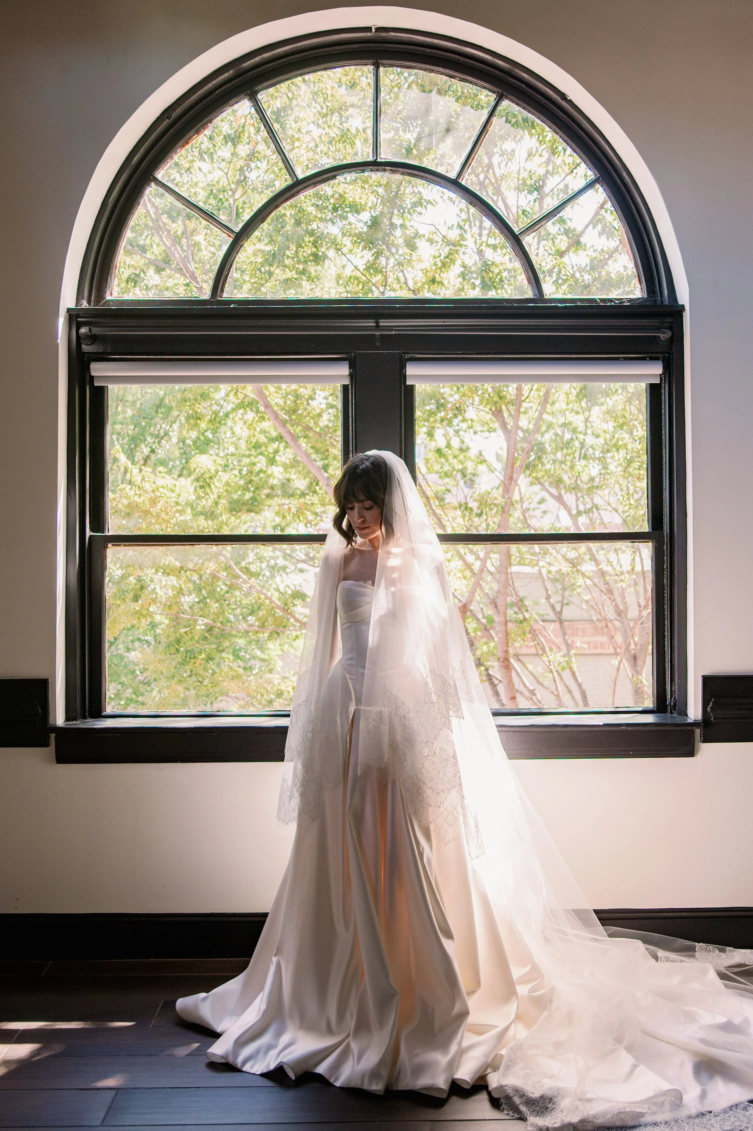 Bride in a satin wedding gown and cathedral veil standing in front of the arched window at Market Hall in Raleigh