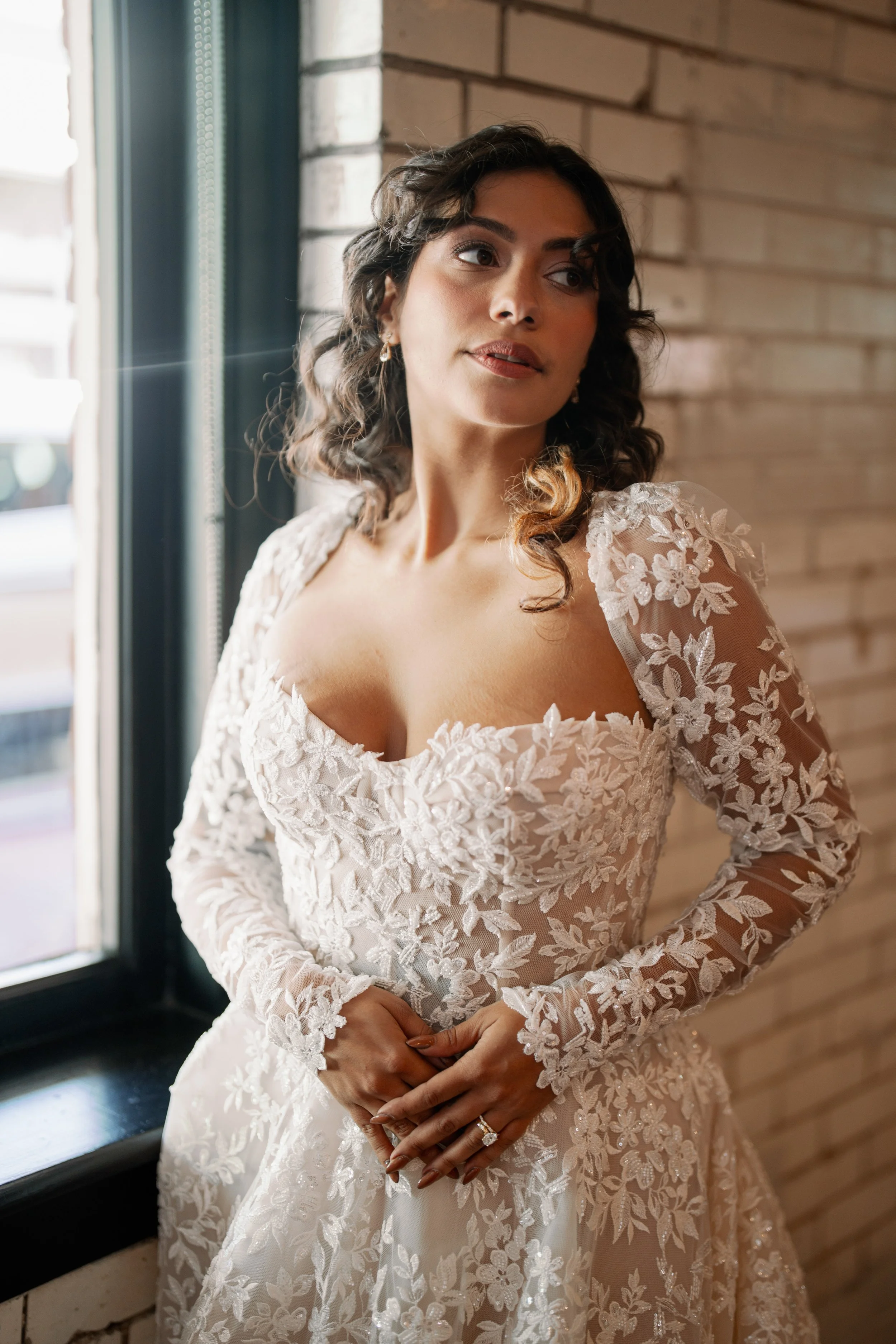 Bride in a long sleeve floral lace gown standing by a window at Market Hall in Raleigh during bridal portraits