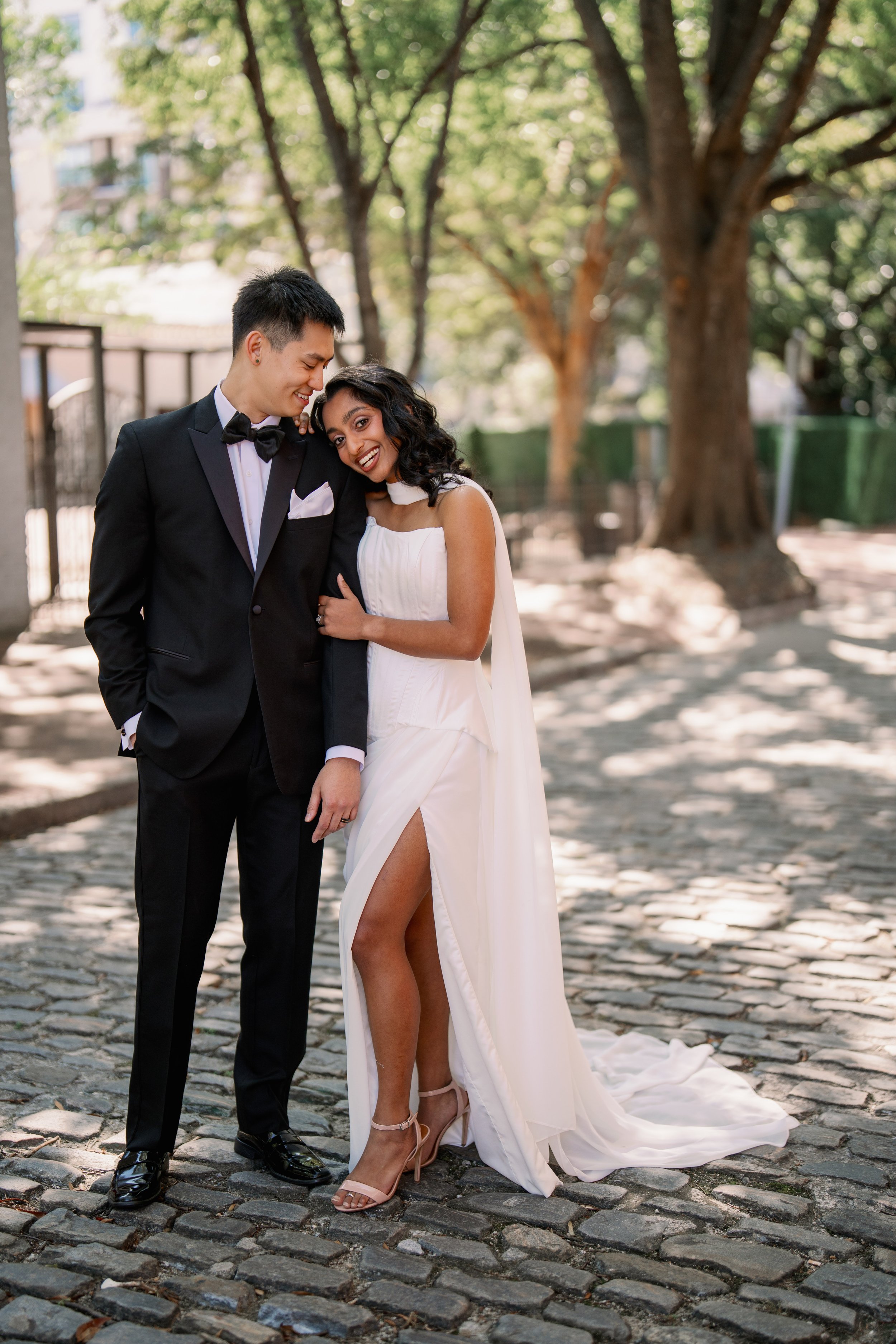 Bride leaning on groom’s shoulder while walking the cobblestone street by Market Hall in Raleigh