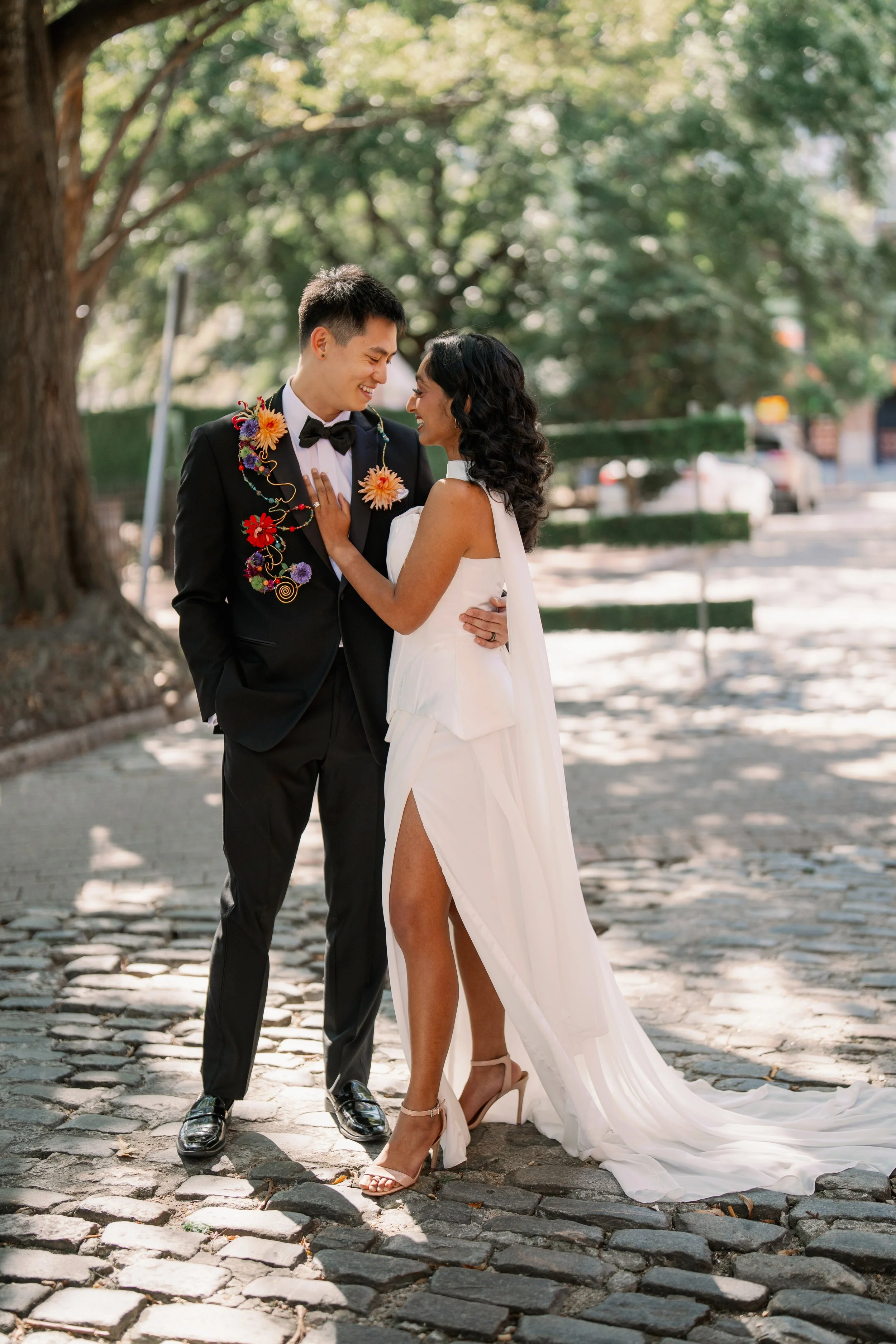 Bride and groom smiling at each other on the cobblestone street outside Market Hall in downtown Raleigh with colorful floral details on the groom’s suit