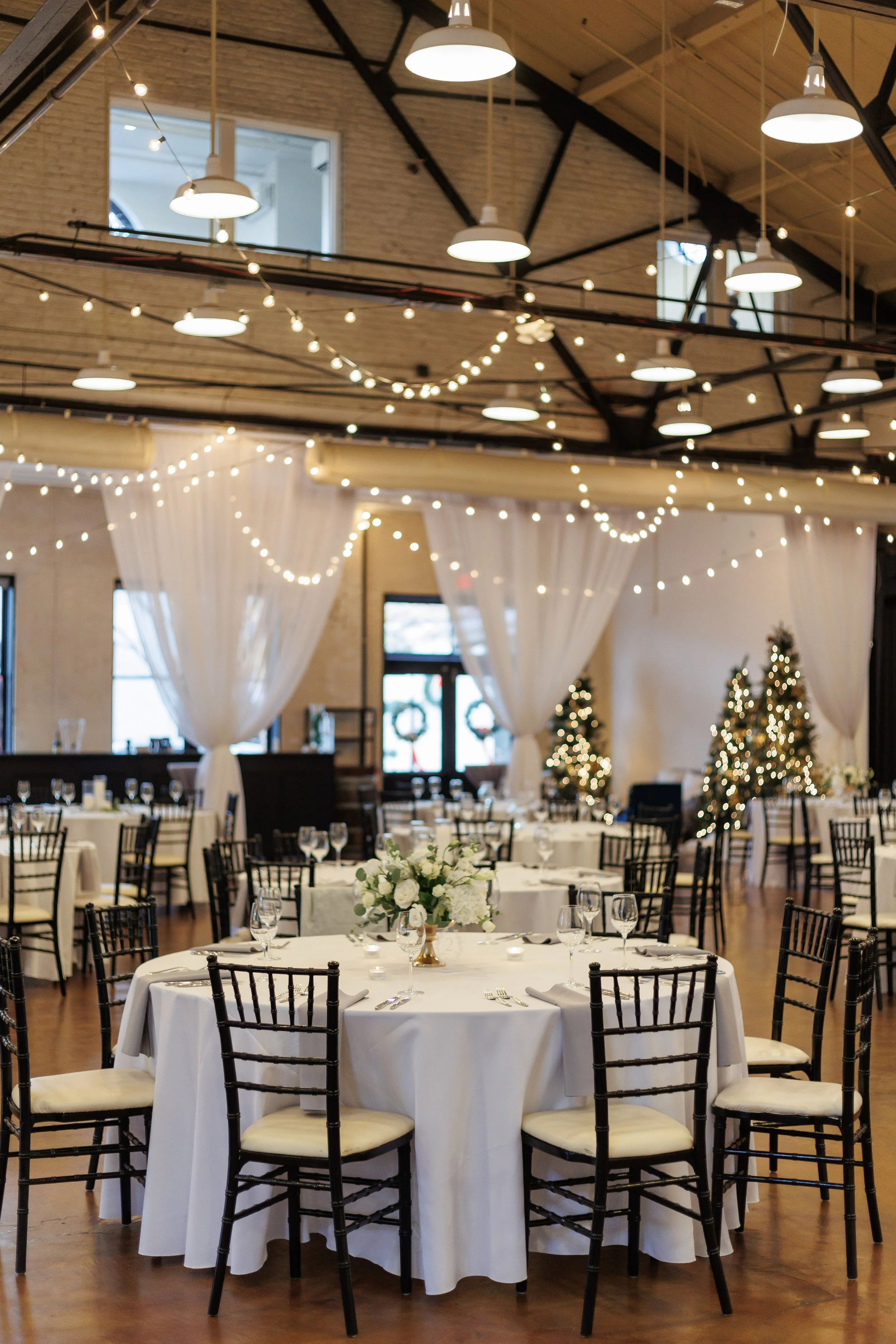 Soft white draping and warm string lights decorate the reception space at Market Hall with round tables and white floral centerpieces.