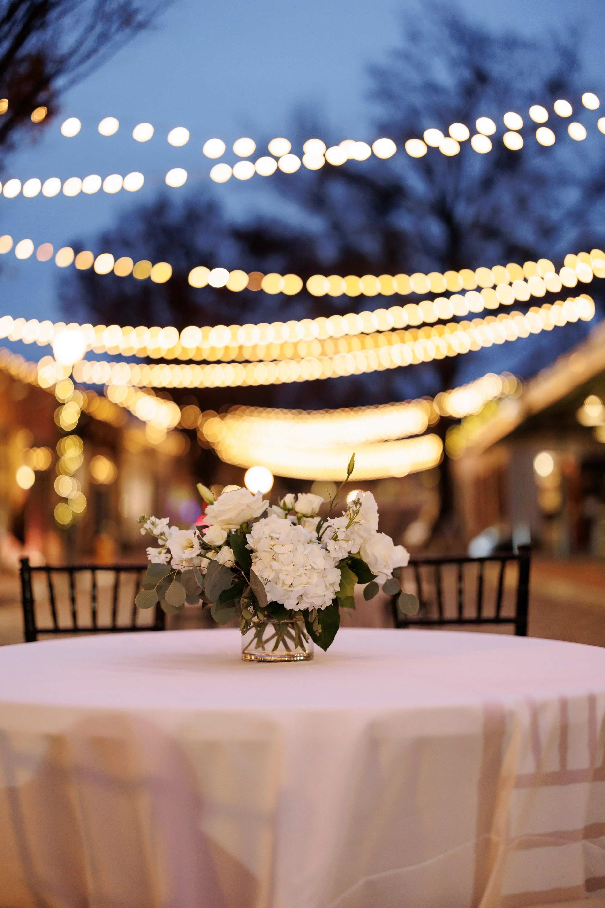 White floral centerpiece on a cocktail table under glowing outdoor string lights at Market Hall in Raleigh.