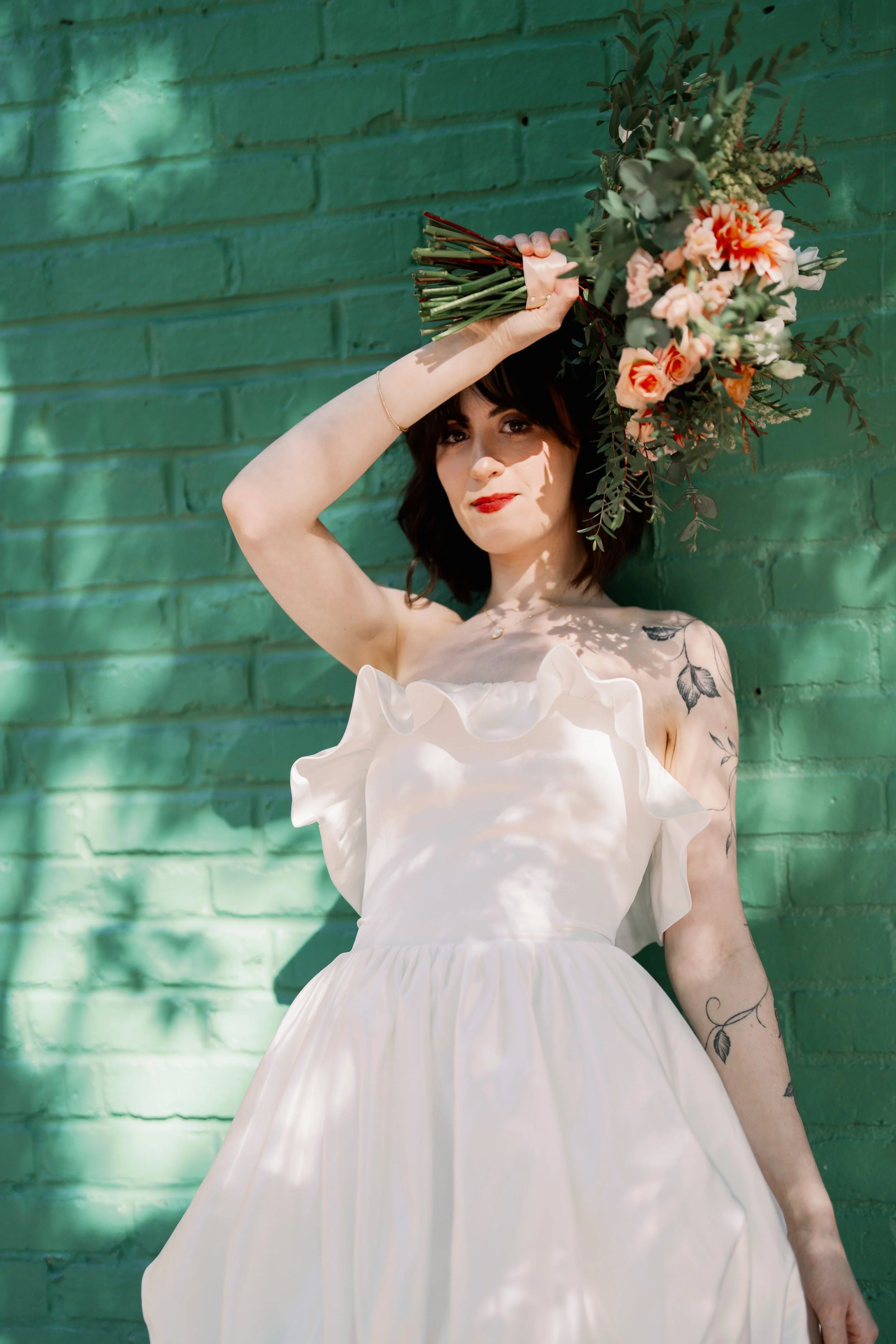 Bride with dark hair in a modern ruffled wedding gown holding a bouquet above her head against a green brick wall at Market Hall in Raleigh North Carolina