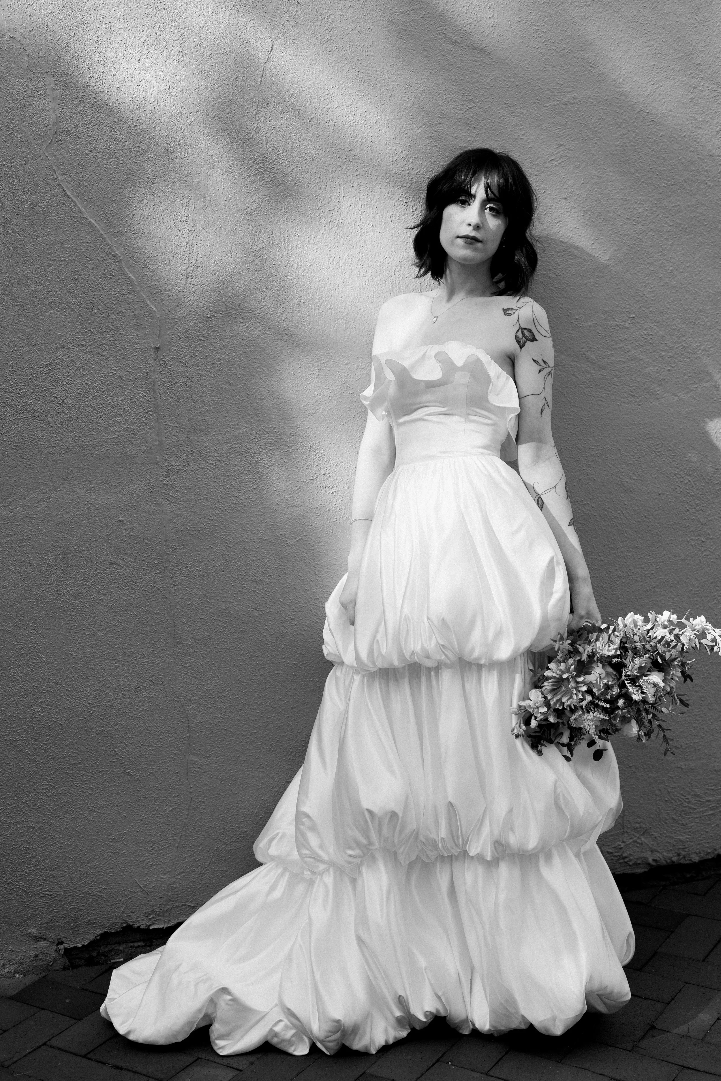 Black and white bridal portrait of a bride in a tiered wedding gown holding flowers beside a textured wall at Market Hall Raleigh