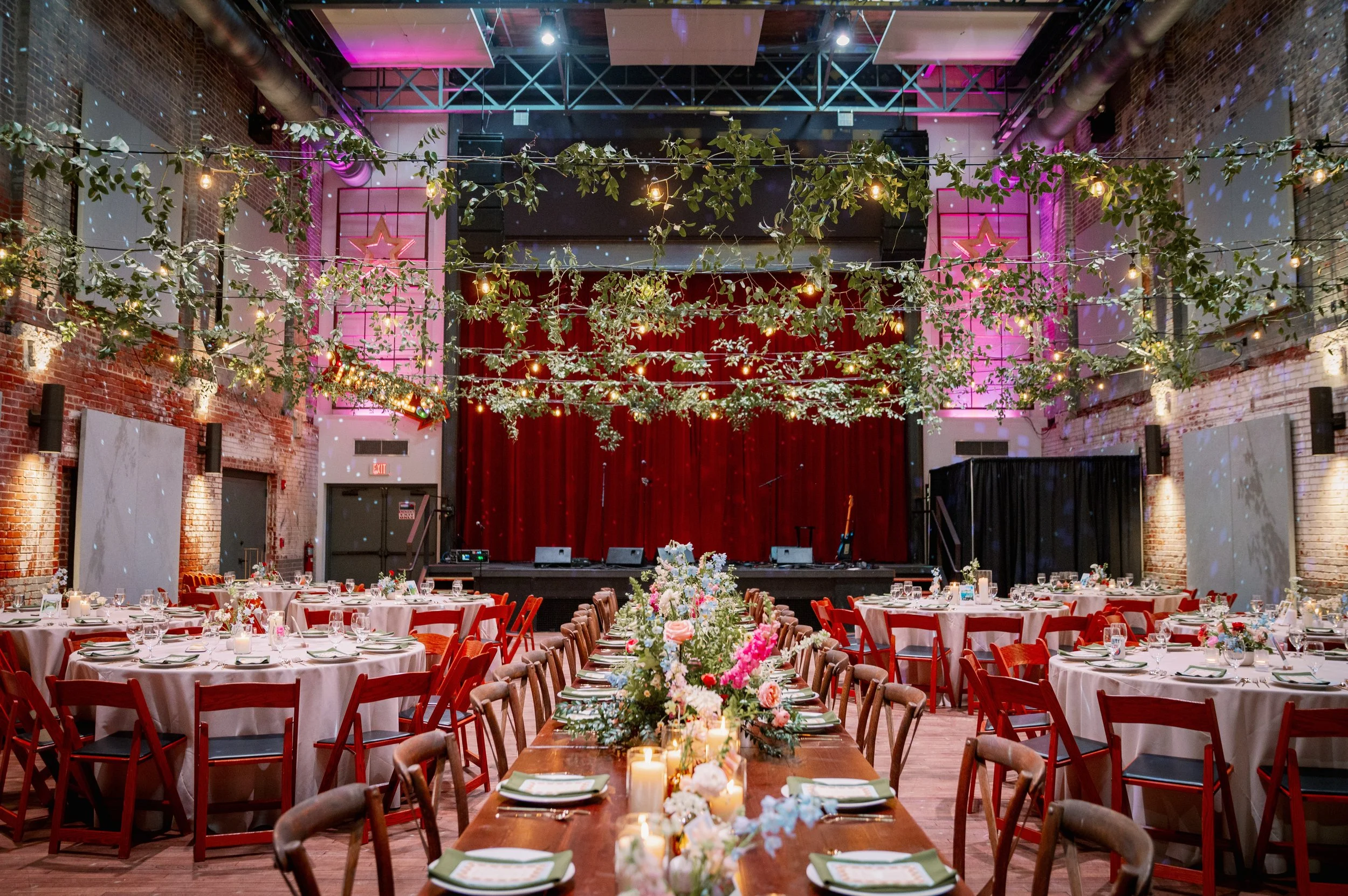  Reception setup at Haw River Ballroom in Saxapahaw North Carolina with long farmhouse tables, round guest tables, greenery overhead, and red stage backdrop 