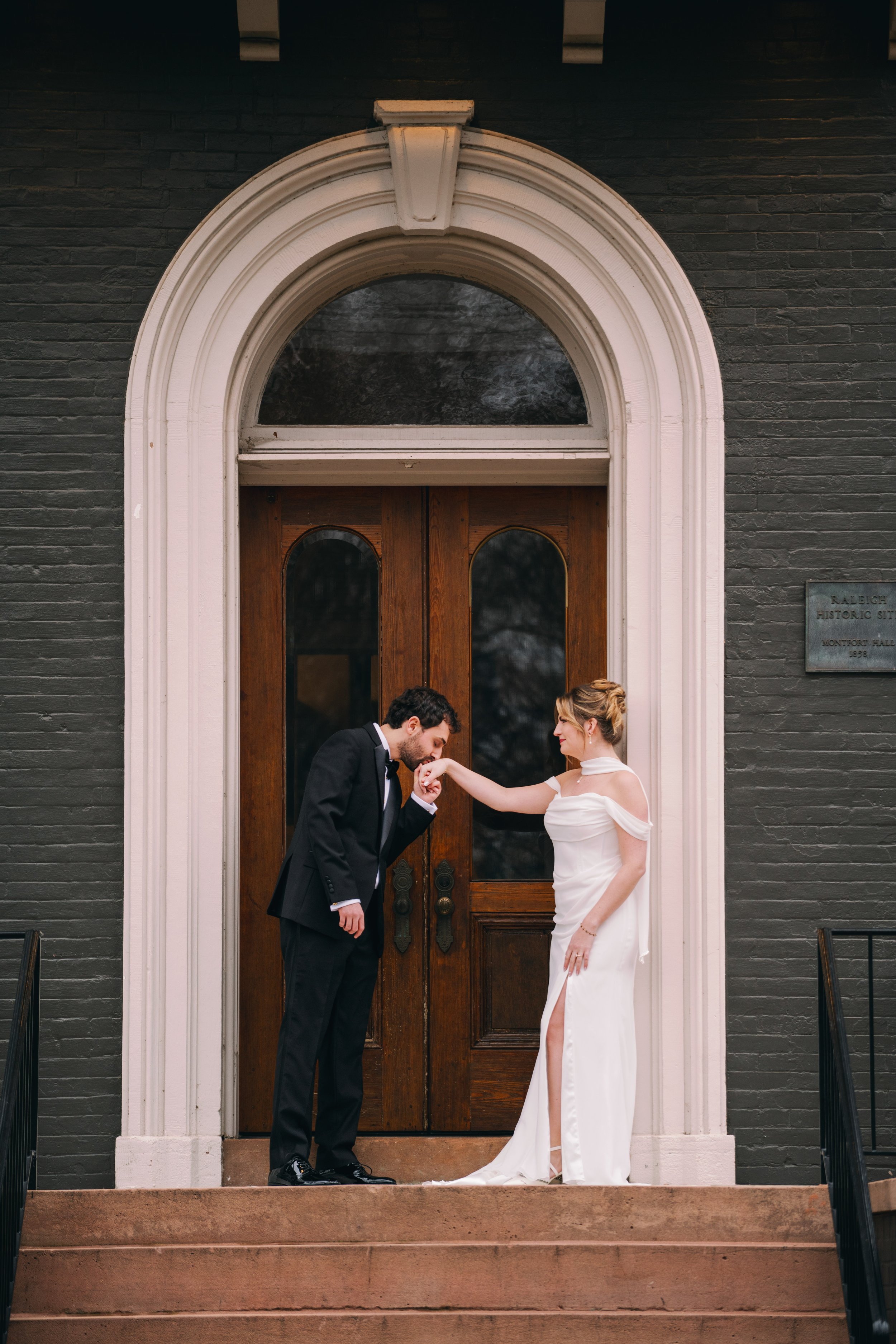  Groom kisses bride's hand on the front steps of Heights House Hotel's arched entryway 
