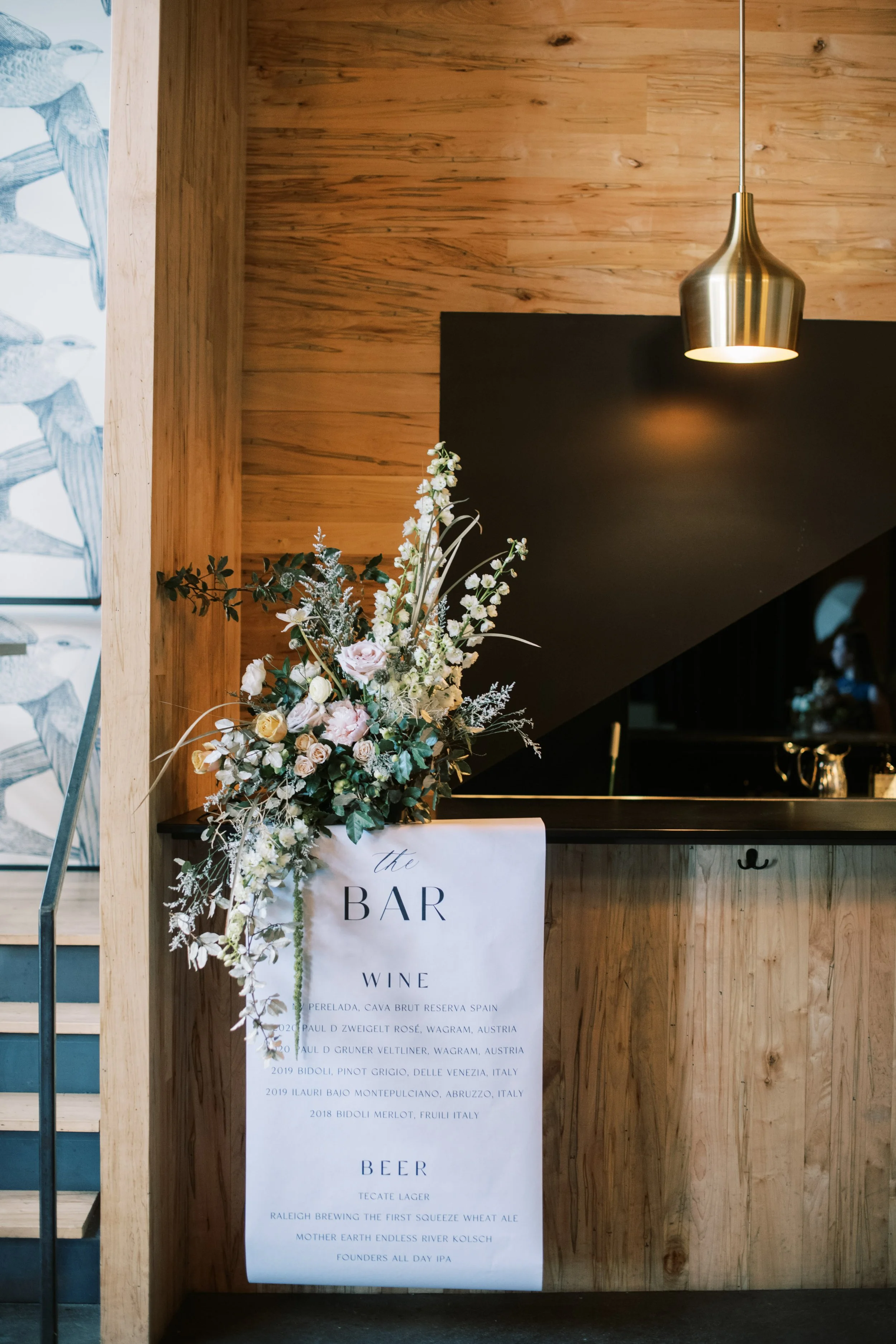 A minimalist bar setup at a Whitaker and Atlantic wedding, with a custom bar menu draped in florals against warm wood walls, blending modern design with natural, refined details.