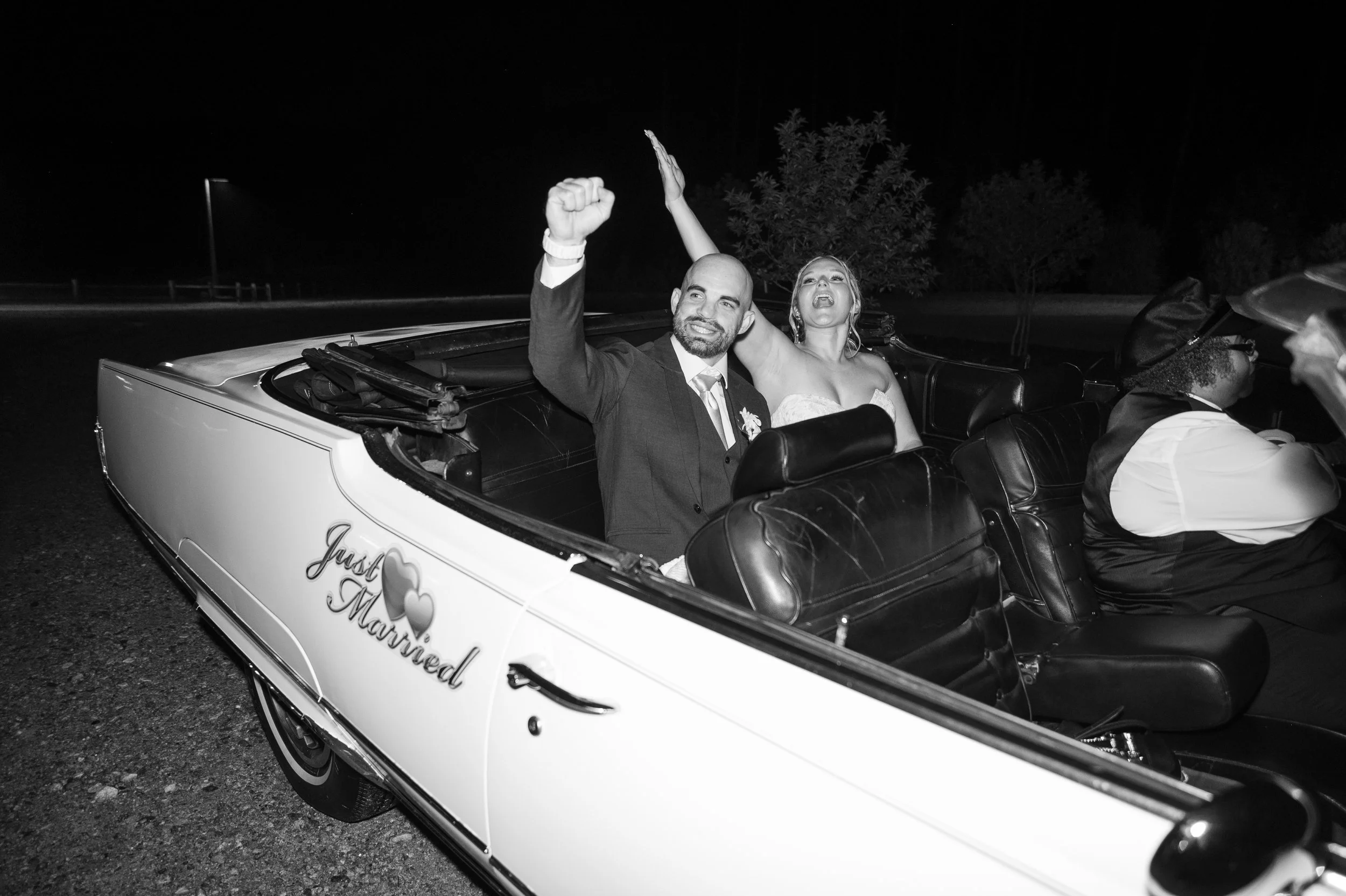 A newlywed couple in a convertible car celebrating their wedding night, with a black and white filter. The groom is smiling with a raised fist, and the bride is making a joyful expression. The side of the car has a sign that reads 'Just Married' with two hearts.