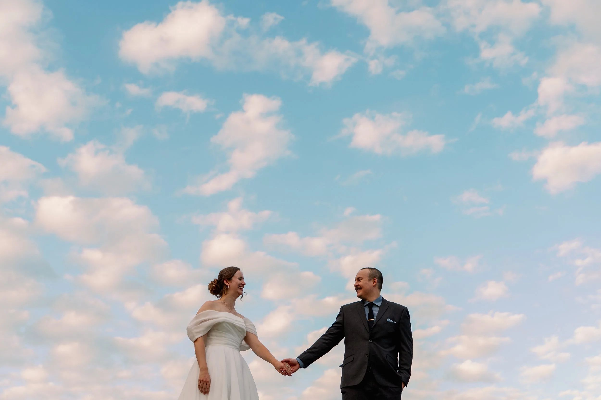 A bride and groom holding hands, smiling at each other outdoors with a sky filled with scattered clouds.