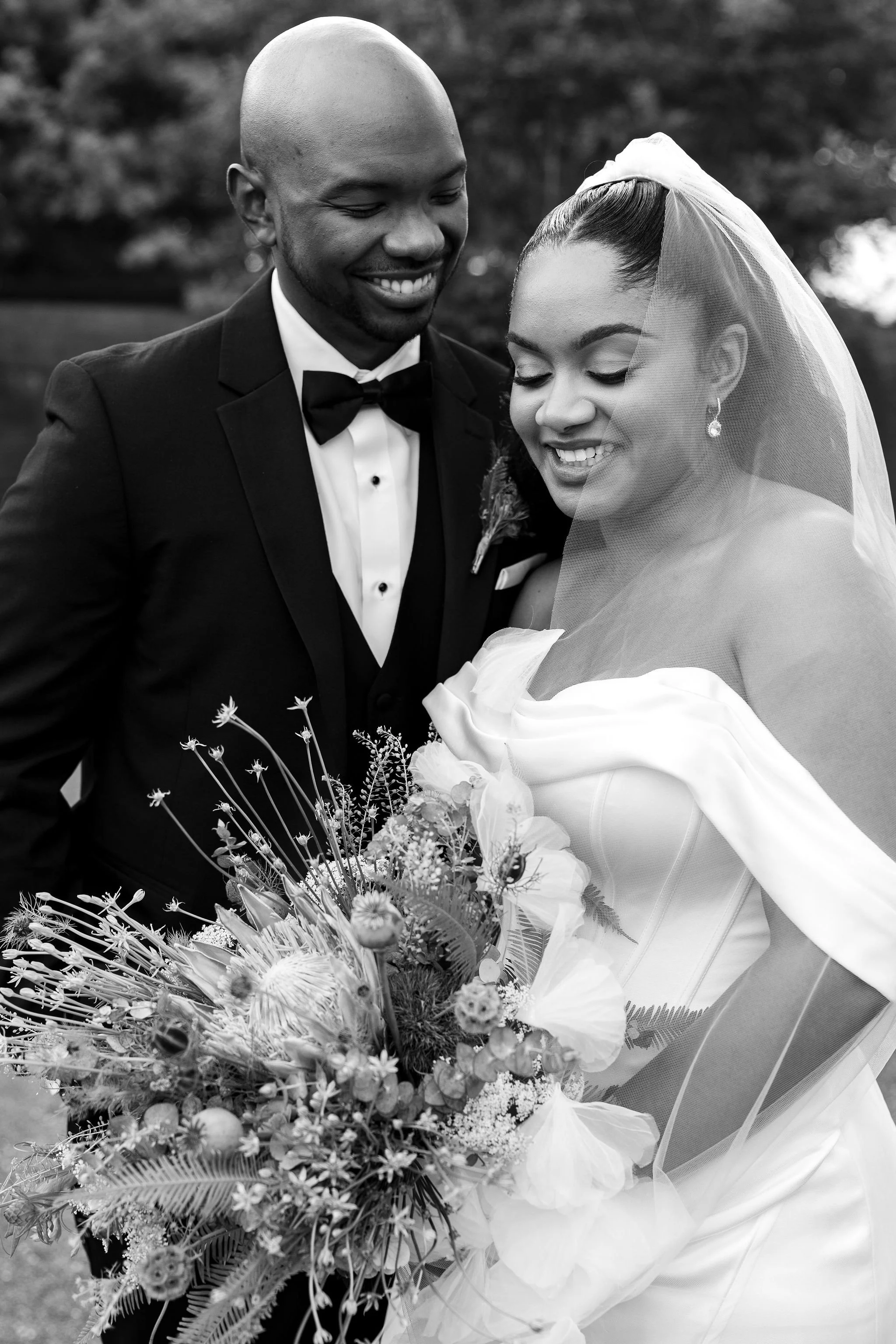 Bride and groom laughing together during outdoor wedding portraits in Raleigh NC, bride holding oversized wildflower bouquet