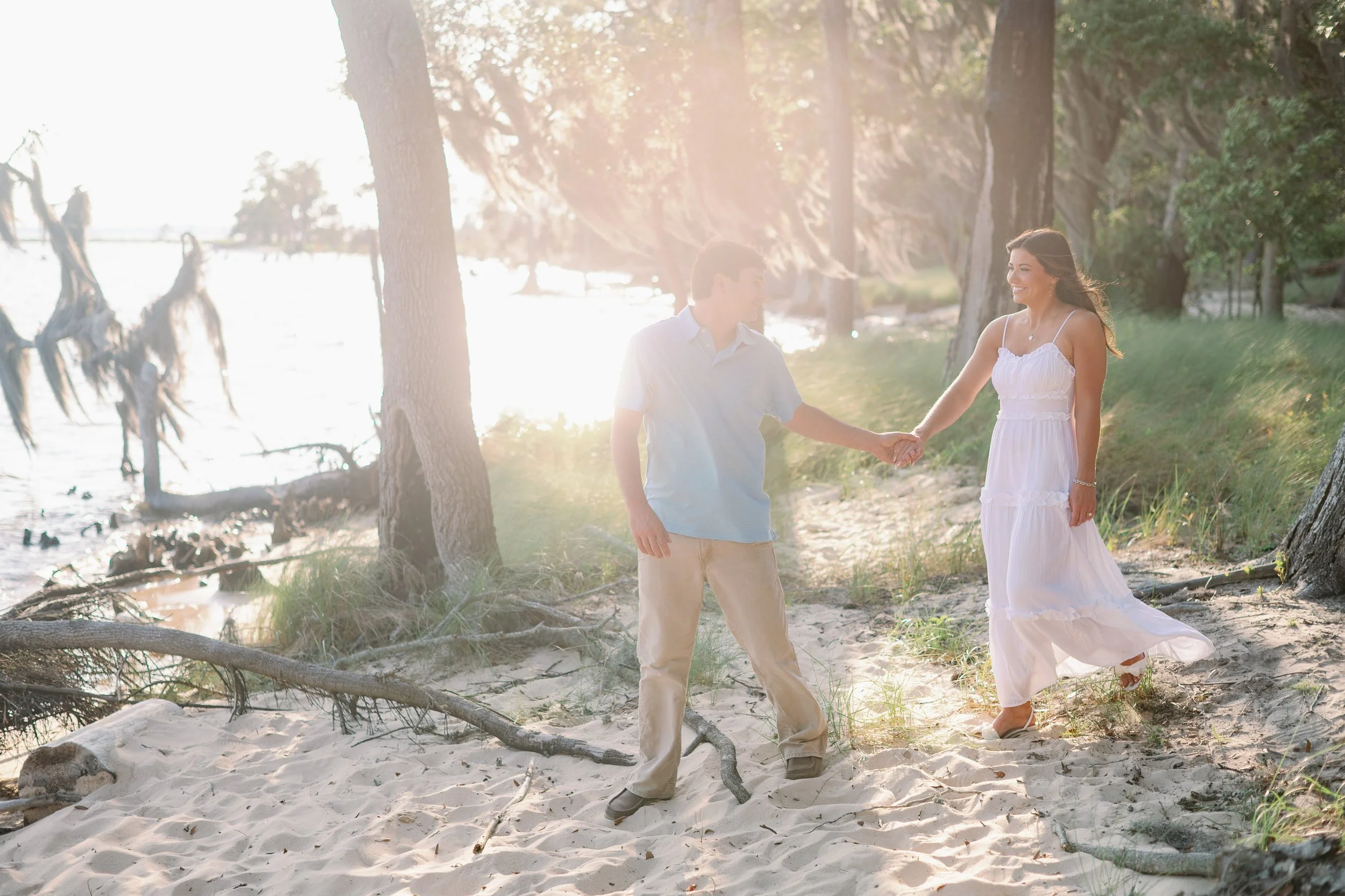 Engagement couple walking hand in hand along the sandy shoreline at Goose Creek State Park in Washington NC with golden backlight through the trees