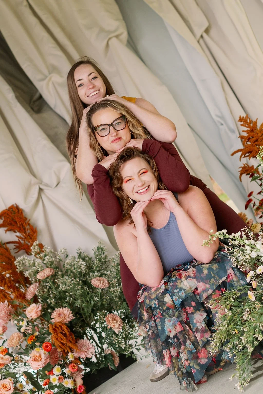 Three women are stacked in a playful pose surrounded by colorful flowers, smiling.