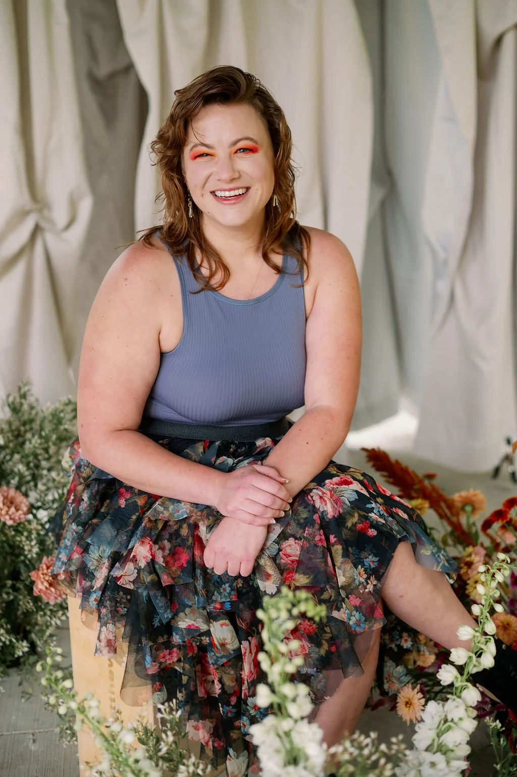 A cheerful woman with wavy brown hair, wearing a blue sleeveless top and a floral skirt, sitting with her hands resting on her lap, surrounded by flowers and a light-colored fabric backdrop.