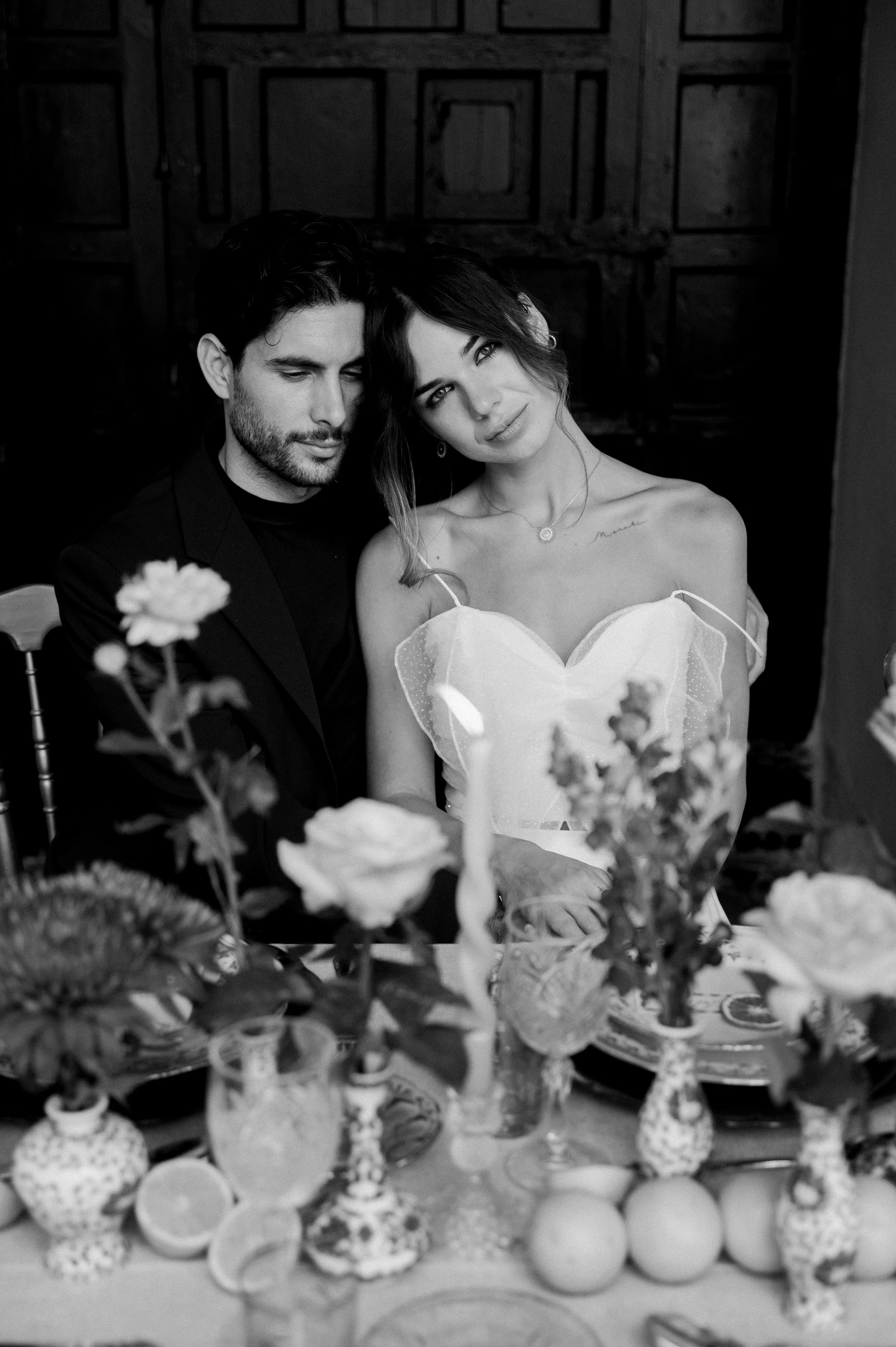 An intimate black and white portrait of a bride and groom seated side by side at a beautifully styled wedding table in Seville. The couple leans into one another surrounded by soft florals, candles, and textured table details, creating a quiet and ro