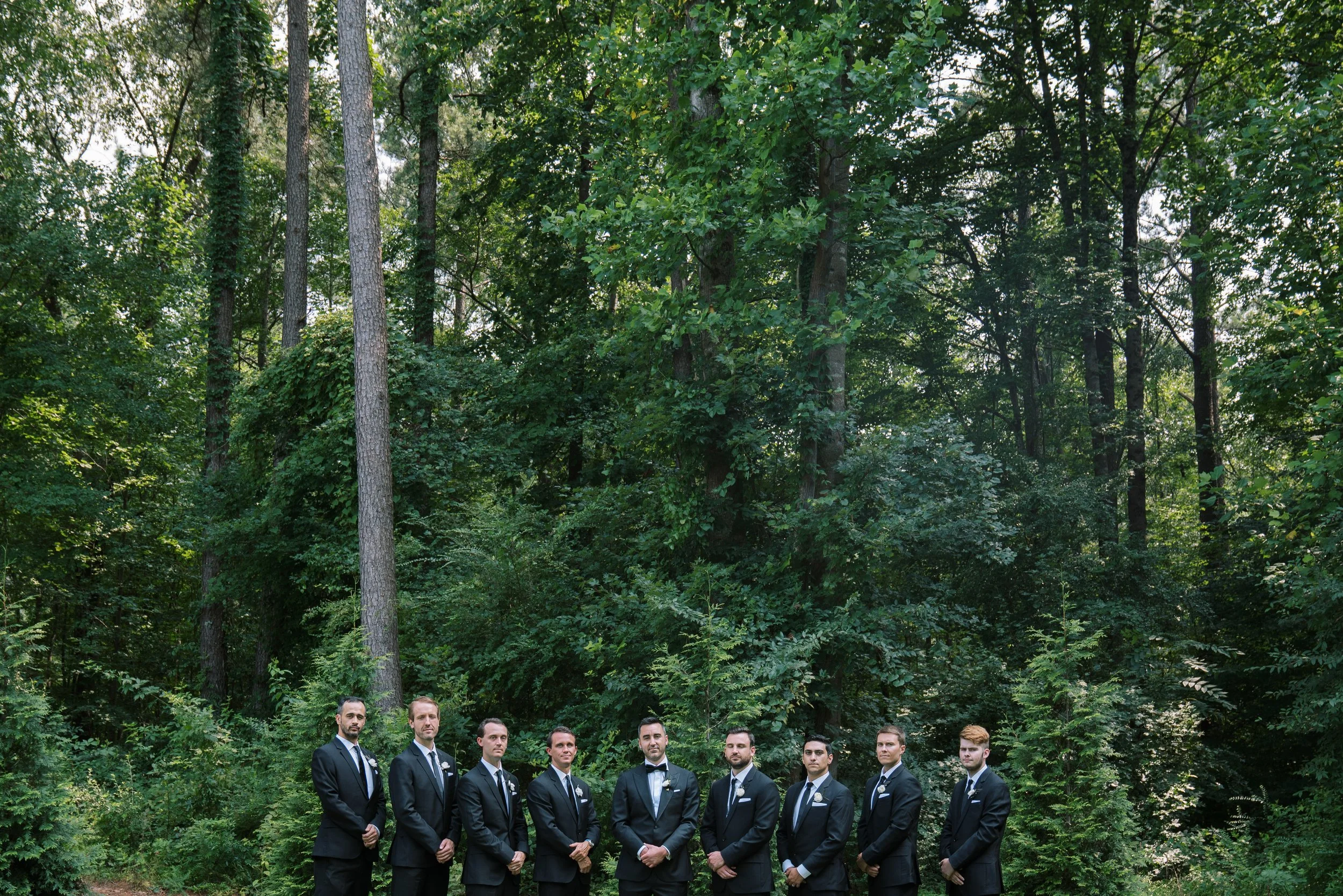 Groomsmen in black tuxedos pose together in a wooded outdoor setting at The Bradford wedding venue in North Carolina.