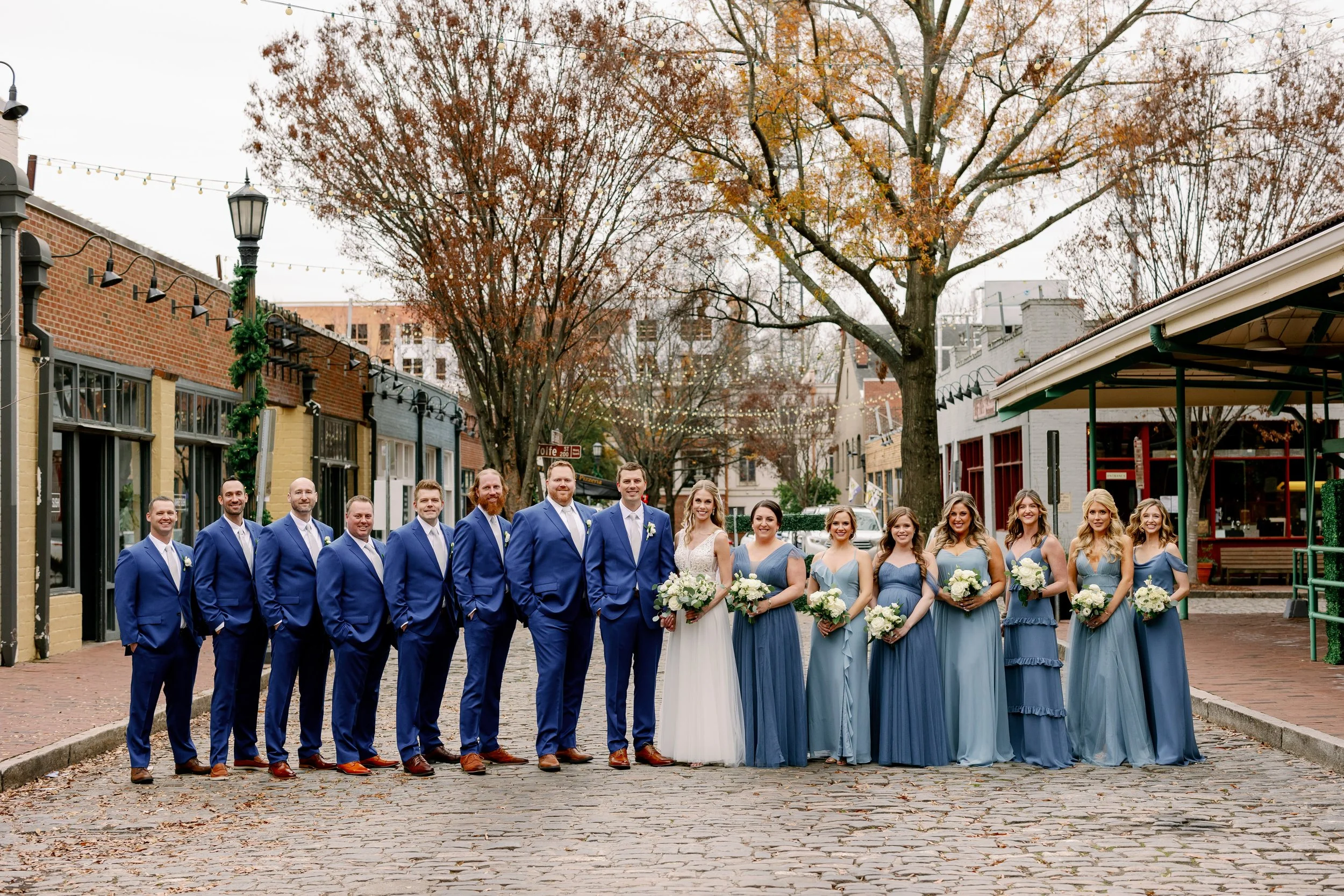 Wedding party standing on the cobblestone street in City Market Raleigh beside Market Hall with groomsmen in blue suits and bridesmaids in blue dresses holding white bouquets.