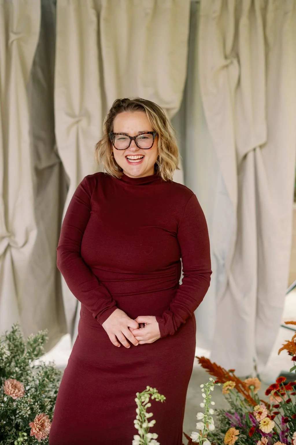 A woman with shoulder-length blonde hair, wearing glasses and a burgundy turtleneck dress, smiling and standing in front of a beige curtain with flowers around her.