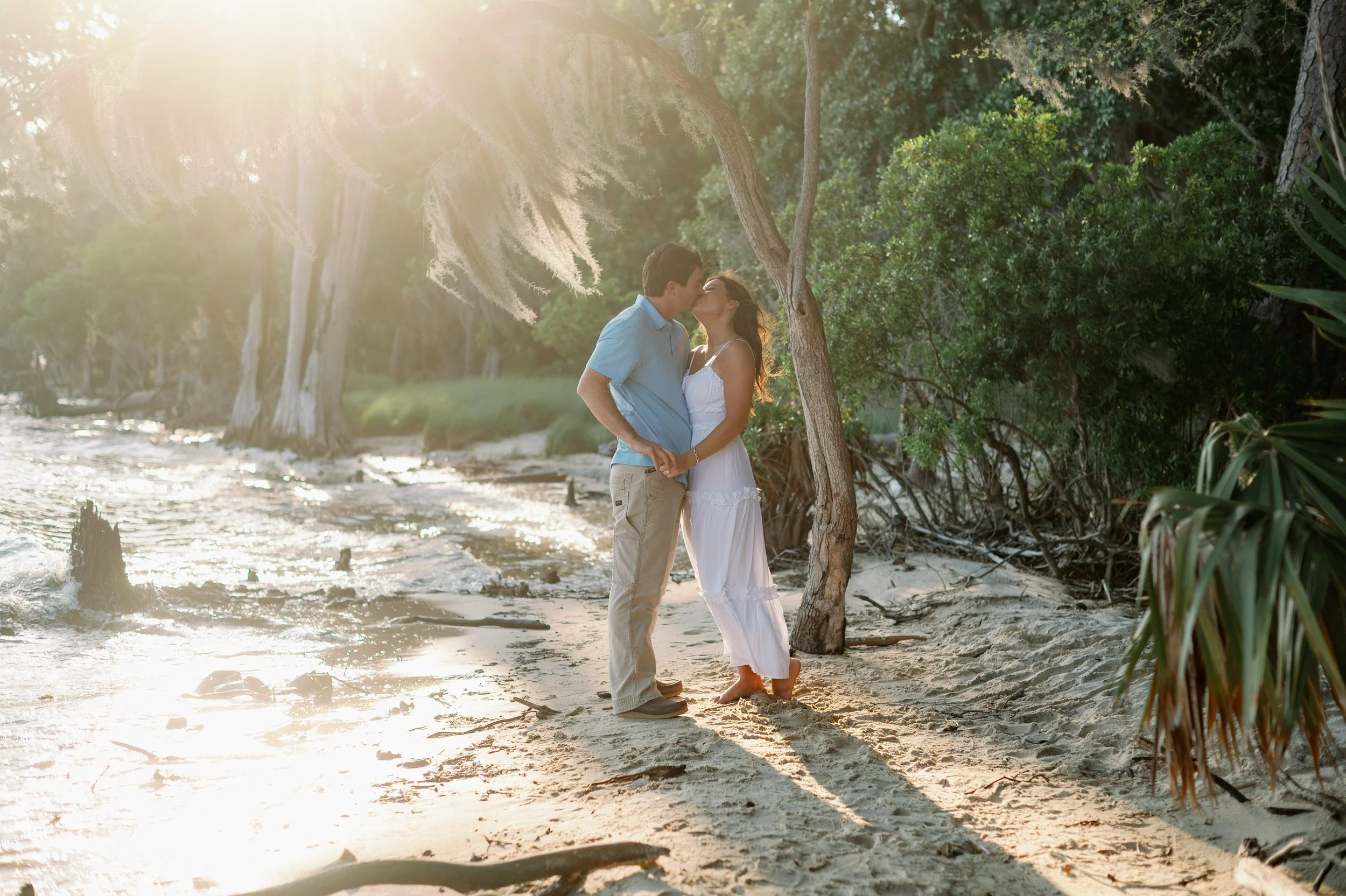 Couple sharing a kiss on the Pamlico River shoreline surrounded by golden backlight and Spanish moss draped trees at Goose Creek State Park in Washington NC