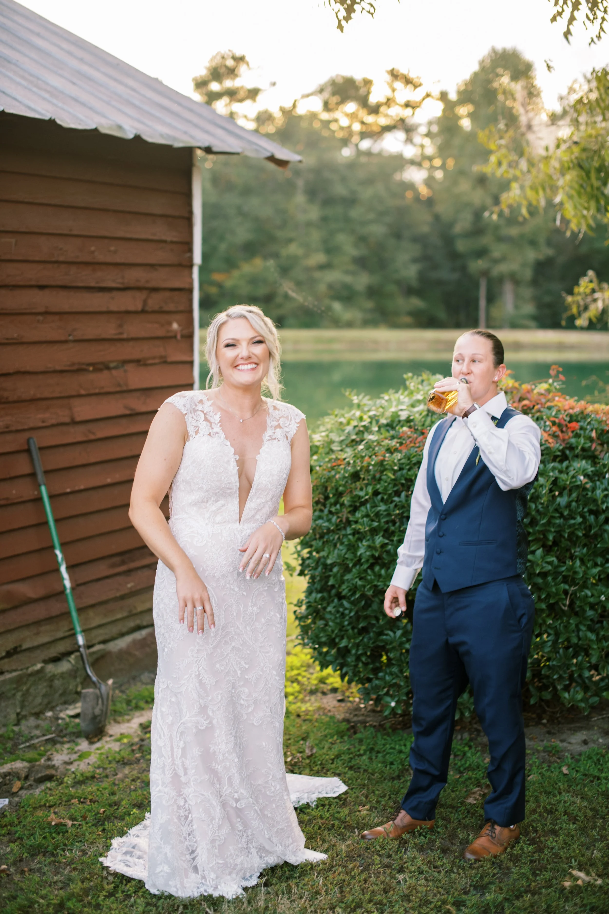 A joyful couple — one bride in a deep-V lace gown and her wife in a navy suit — share a candid laugh together outdoors at a rustic farm wedding venue in the Raleigh, NC area.