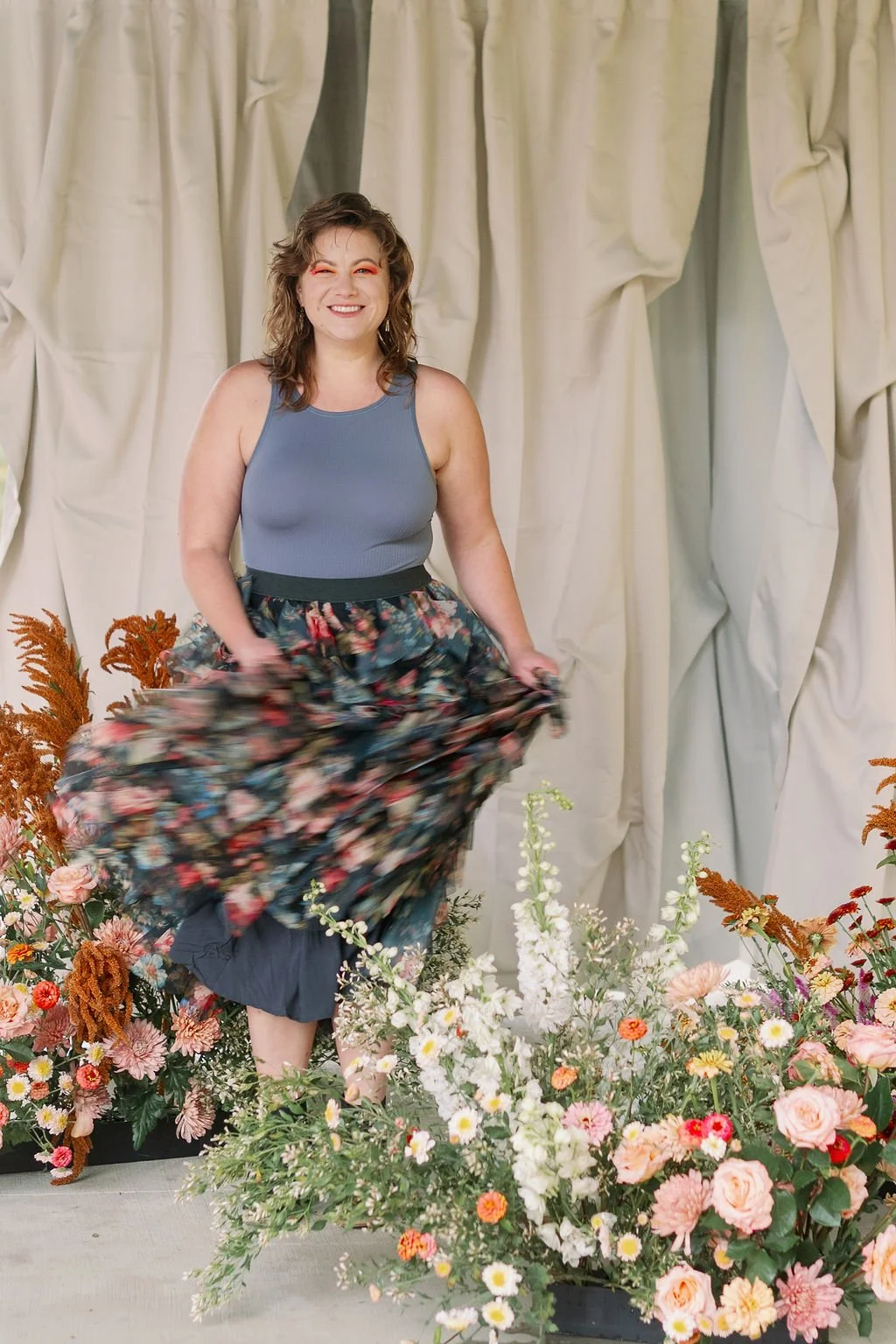 A woman smiling and twirling in front of cream-colored curtains, surrounded by colorful flowers.