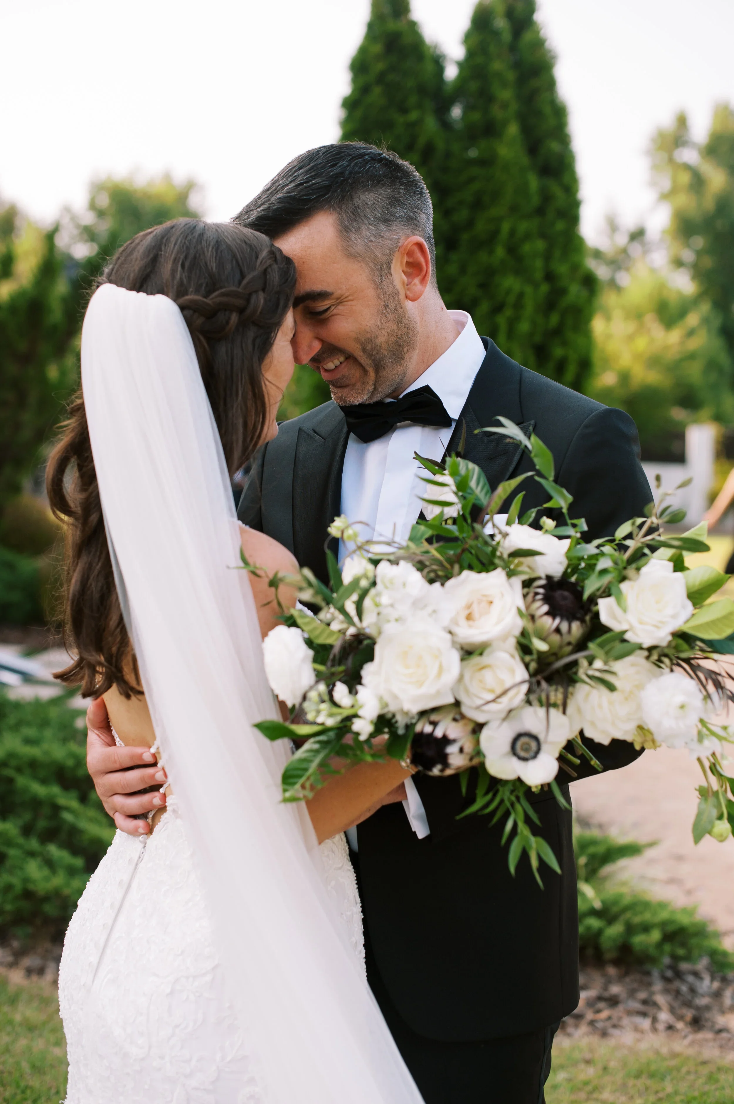 An intimate portrait of the couple during golden hour at The Bradford, tucked into the greenery just outside the venue. They’re pressed forehead to forehead, laughing softly, completely absorbed in the moment. The bride holds a lush bouquet of white 
