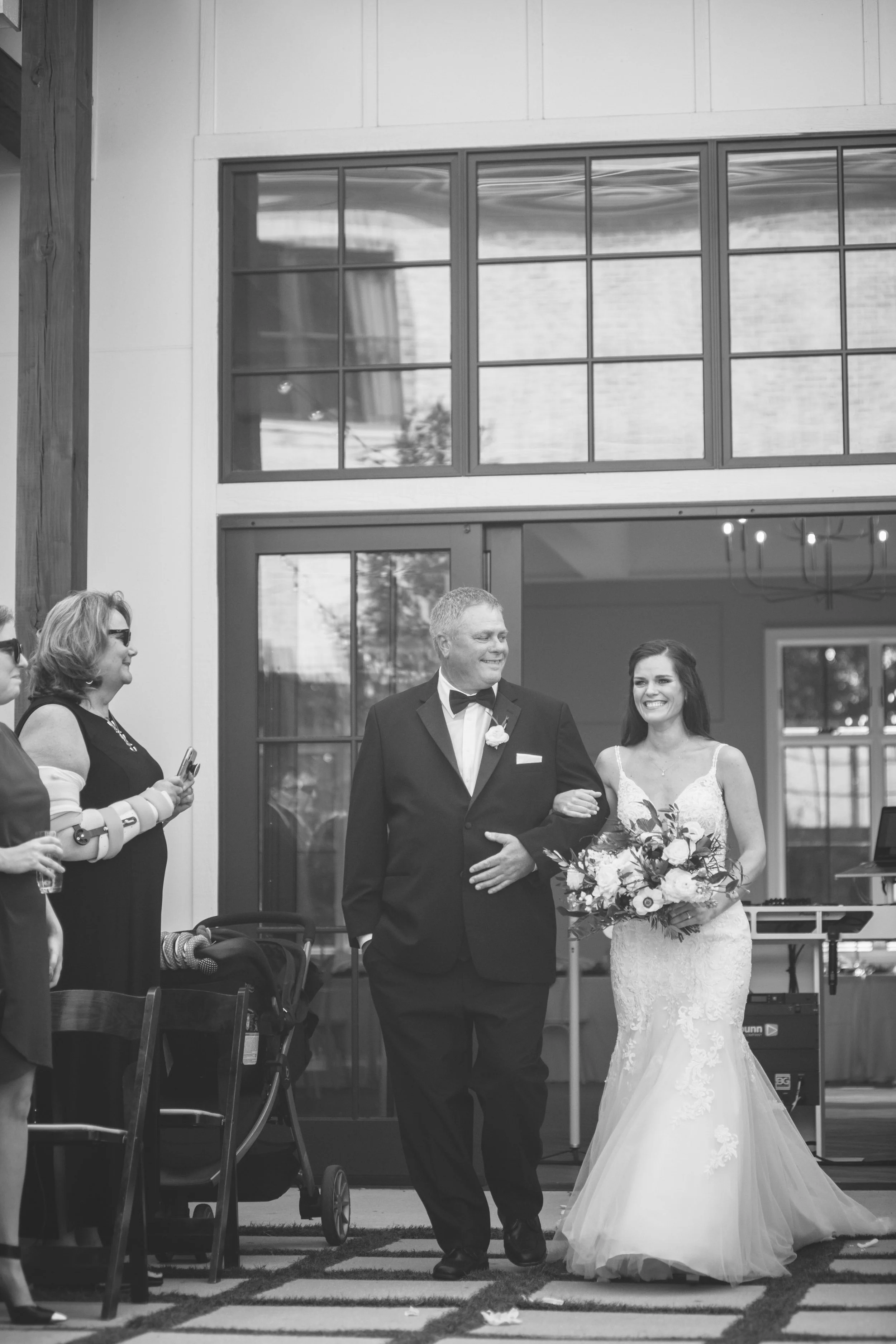 Bride walks down the aisle with her father during an outdoor wedding ceremony at The Bradford wedding venue in North Carolina, captured in black and white.