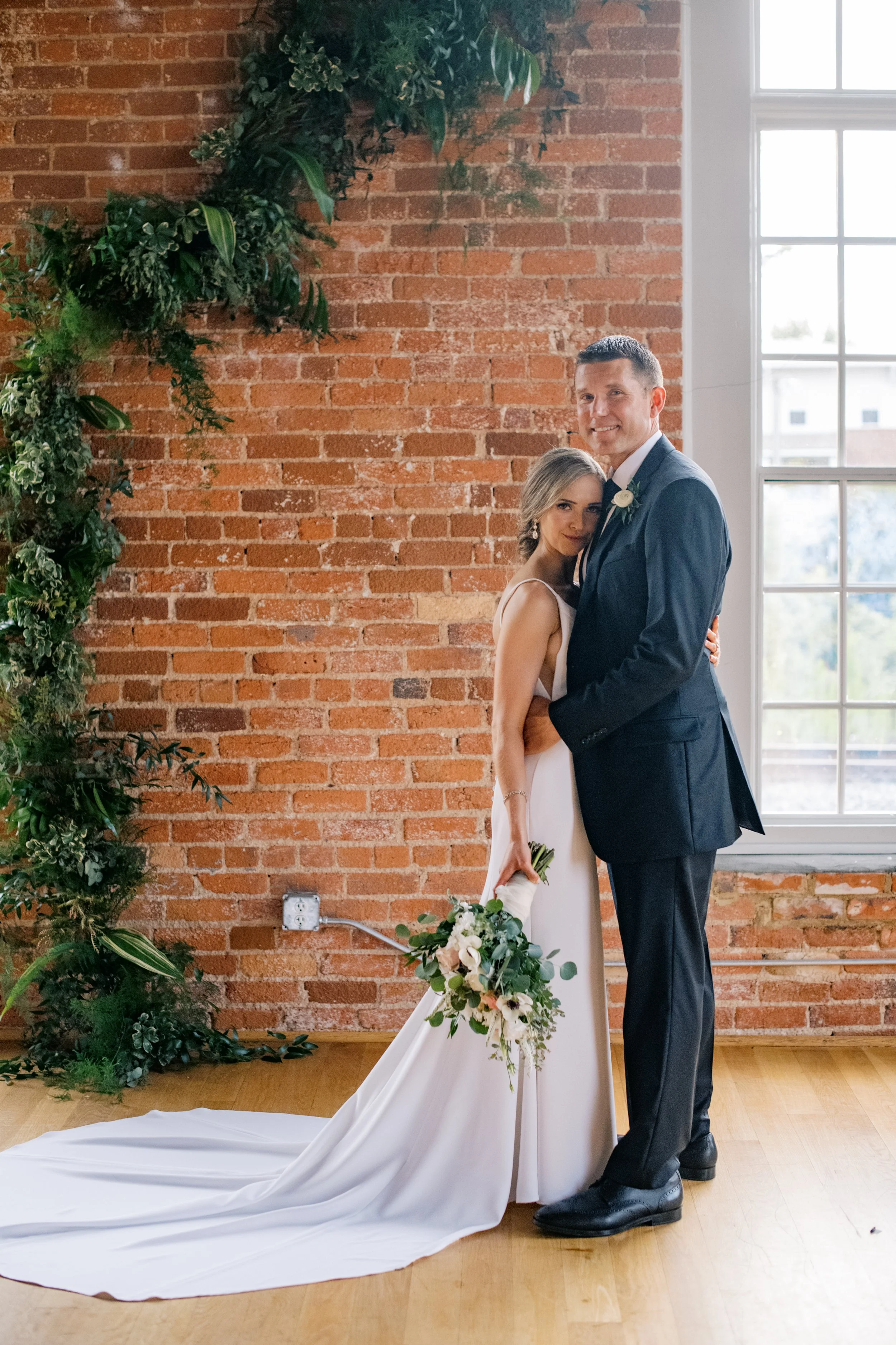 Bride and groom embracing in front of an exposed brick wall with greenery at The Cotton Room wedding venue in Durham, North Carolina.
