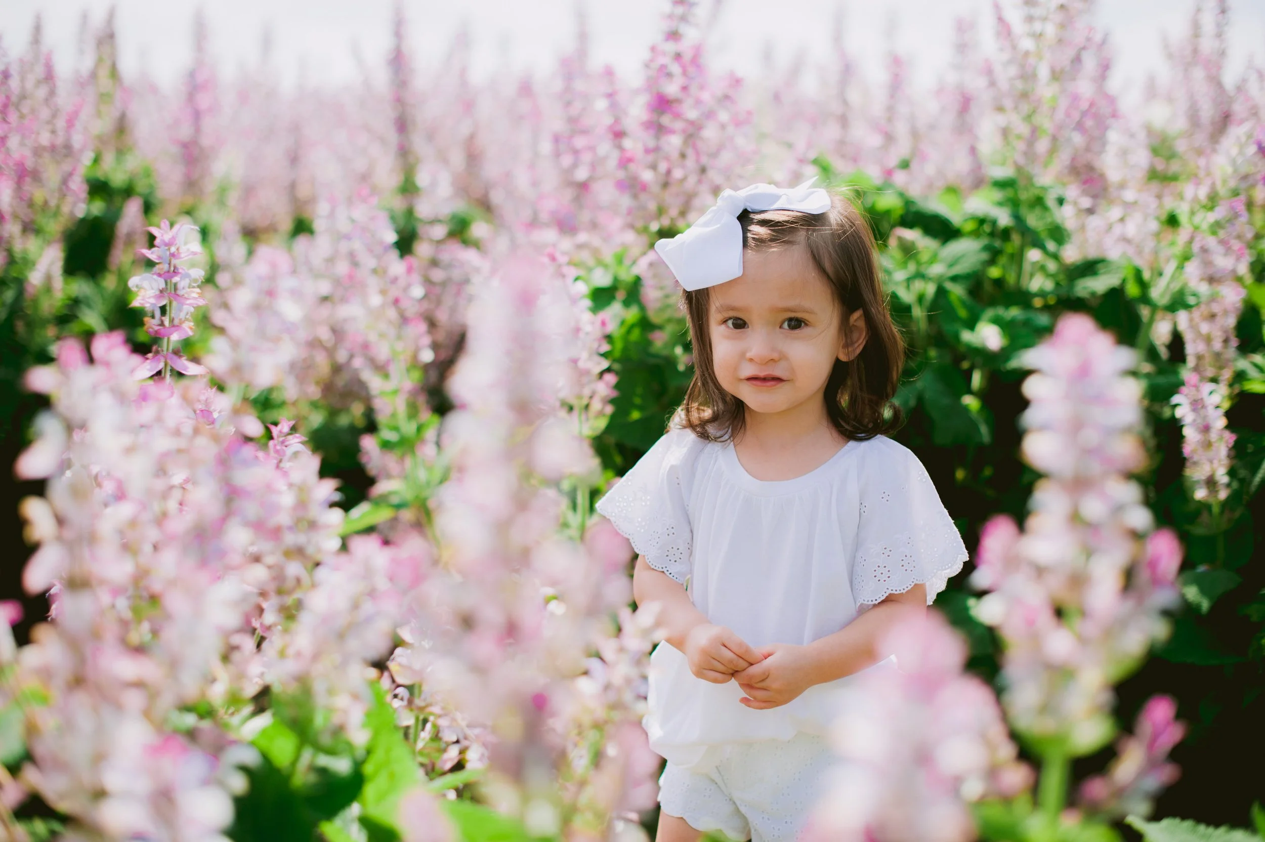 A young girl standing in a field of pink and white flowers, wearing a white dress and a large white bow in her hair, with a slightly distressed or confused expression on her face.