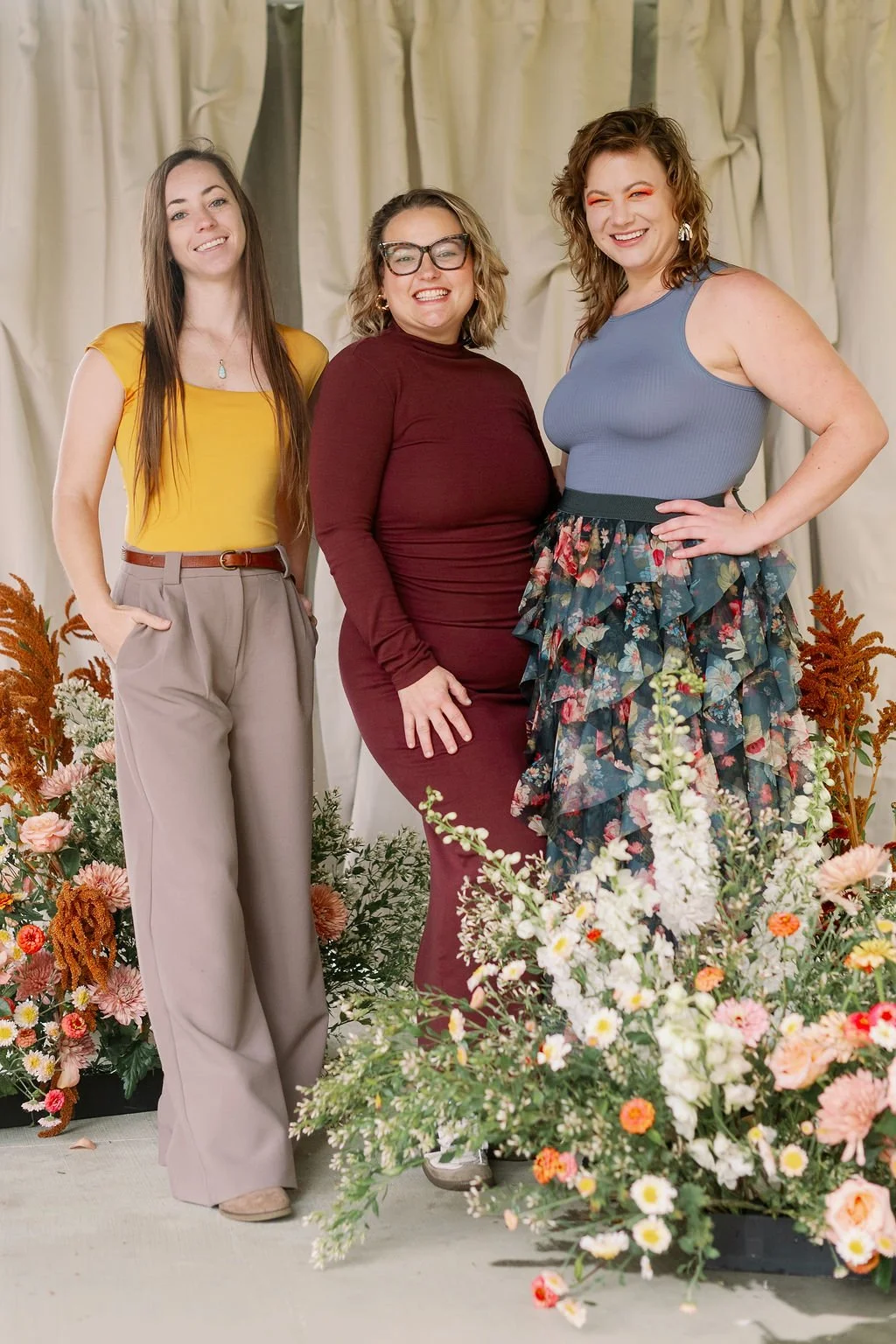 Three women stand together in front of a cream-colored curtain backdrop with floral arrangements at their feet, smiling.