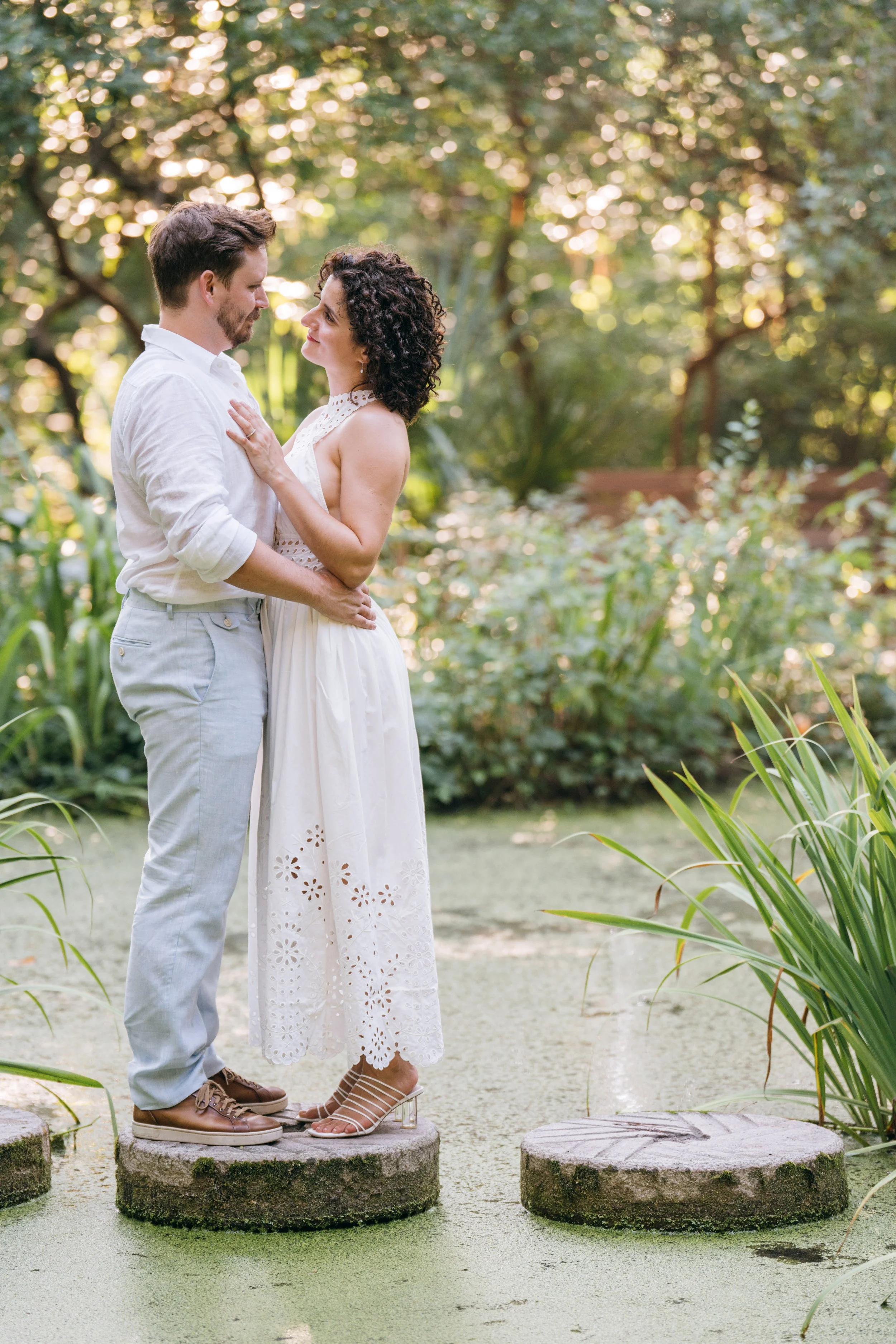 An engaged couple shares a quiet moment while standing on stone steps above a pond at Duke Gardens in Durham, NC. Surrounded by lush greenery and soft natural light, the scene feels intimate and romantic, capturing a candid, documentary style engagem