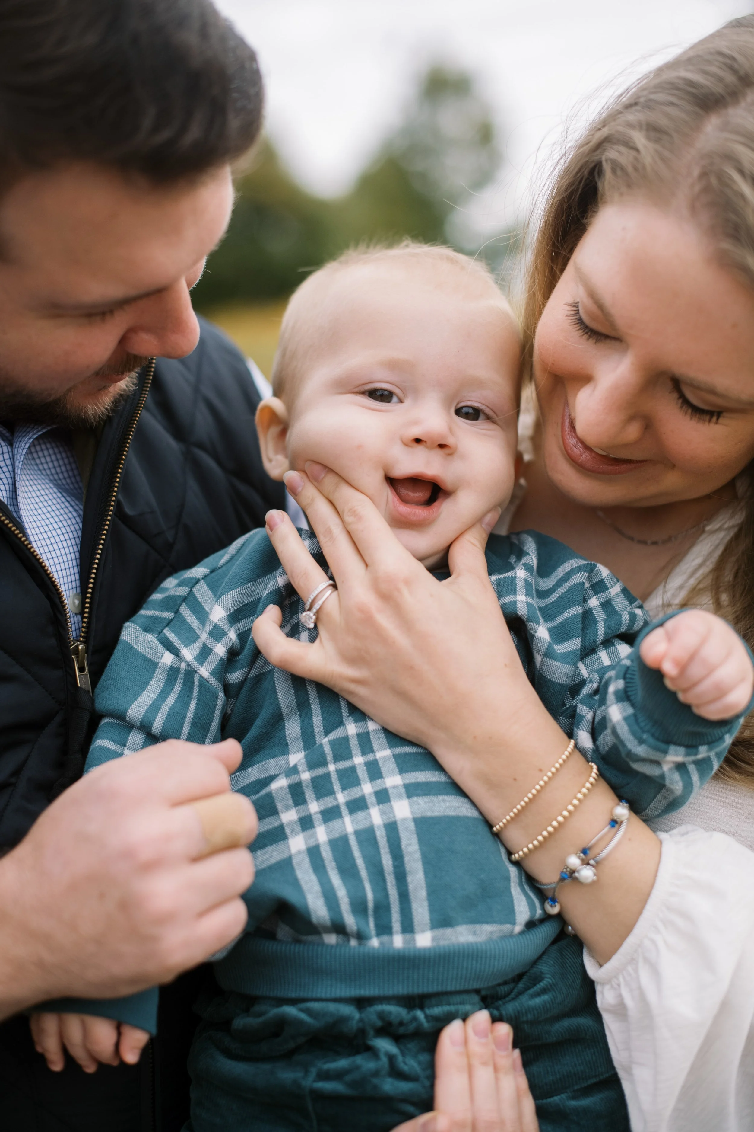 A man and woman holding a happy baby outdoors in a park with trees in the background.