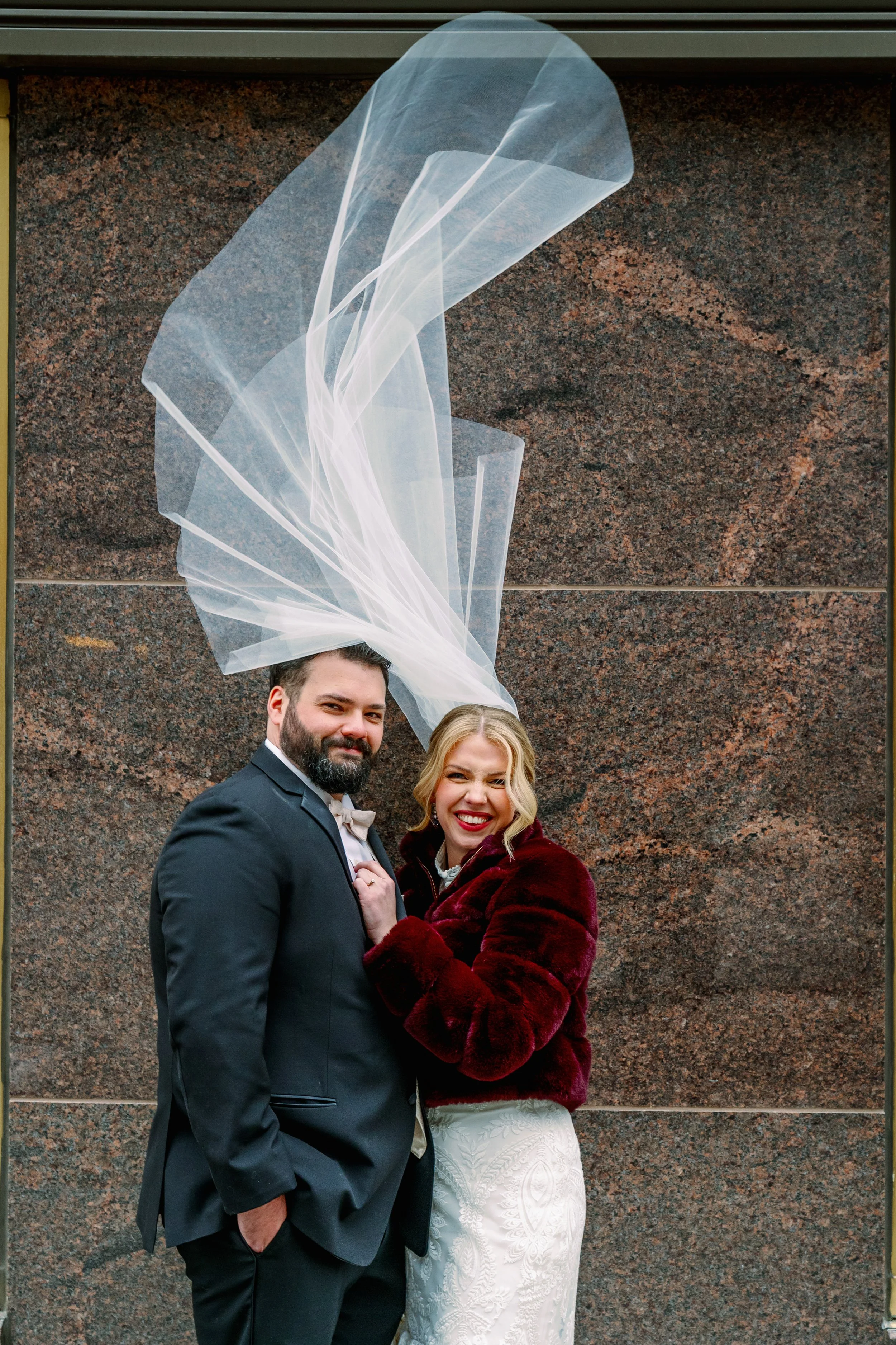 A happy couple poses together during their wedding, standing in front of a brown stone wall. The groom wears a black suit, white shirt, and light-colored bow tie, with his left hand in his pocket. The bride wears a white wedding dress and a red velve