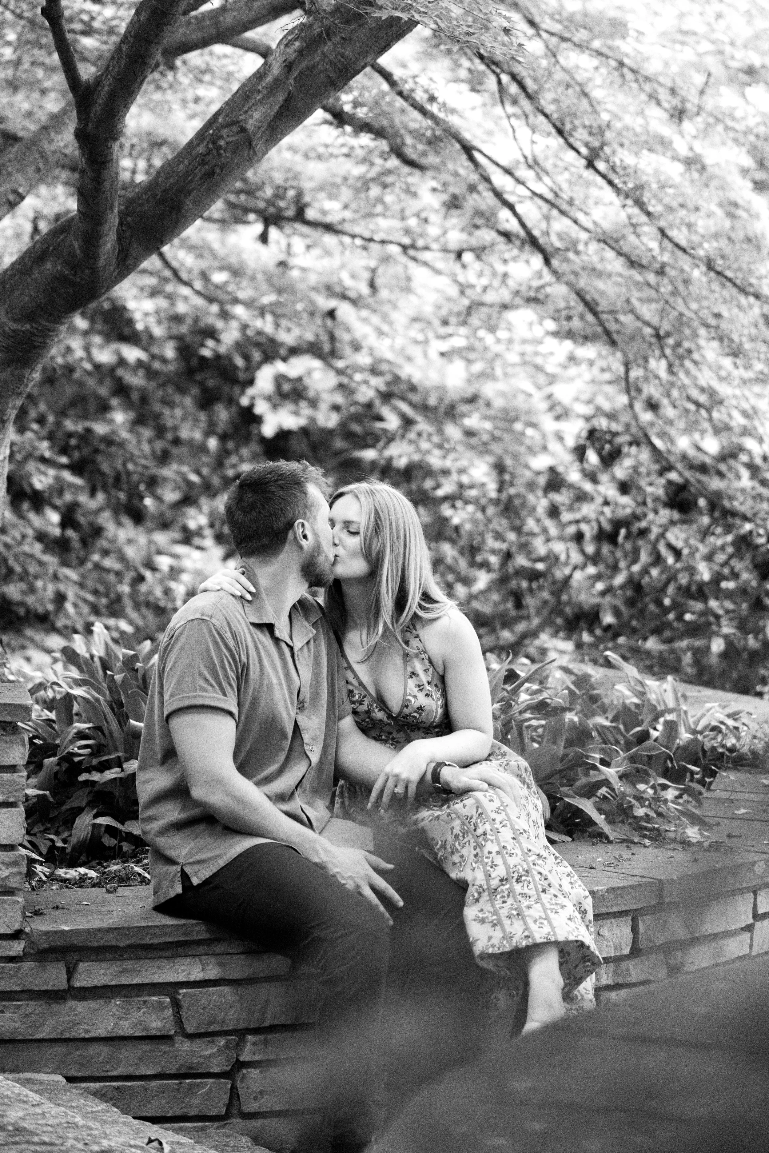 A black and white photo of a couple sharing a quiet kiss during their proposal at Duke Gardens in Durham, North Carolina. They are seated on a stone wall surrounded by lush greenery, with soft natural light filtering through the trees, creating an in