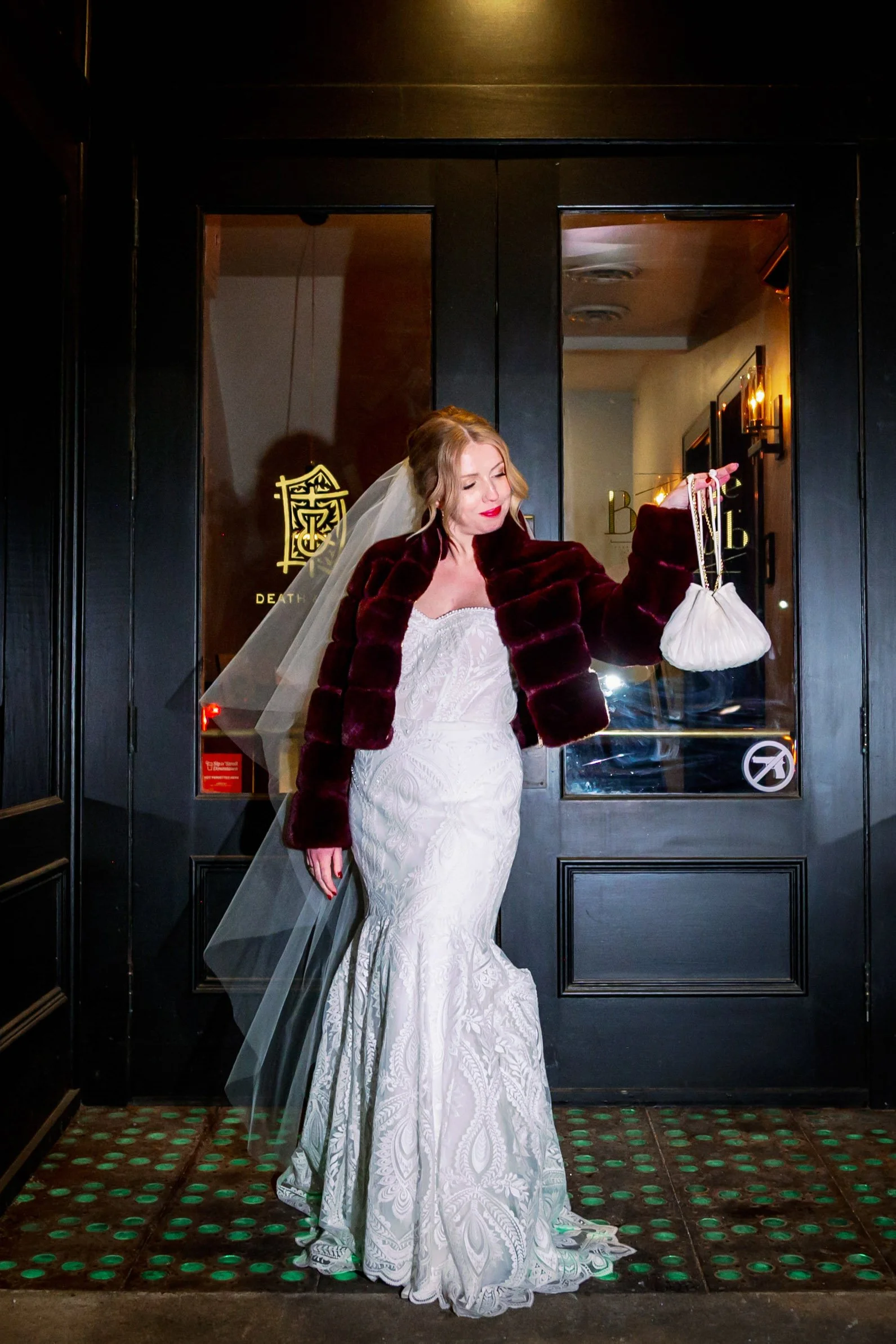 A woman in a white lace wedding dress and burgundy fur jacket stands in front of a black door, holding a small white bag, with a veil on her head and a slight smile.