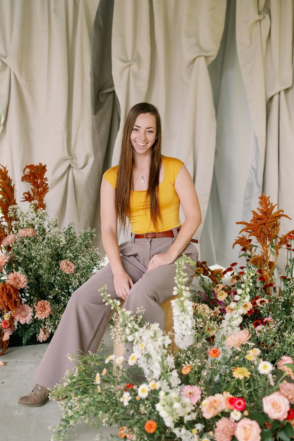 A woman with long brown hair smiling while sitting on a small stool surrounded by various colorful flowers, with a beige curtain backdrop.